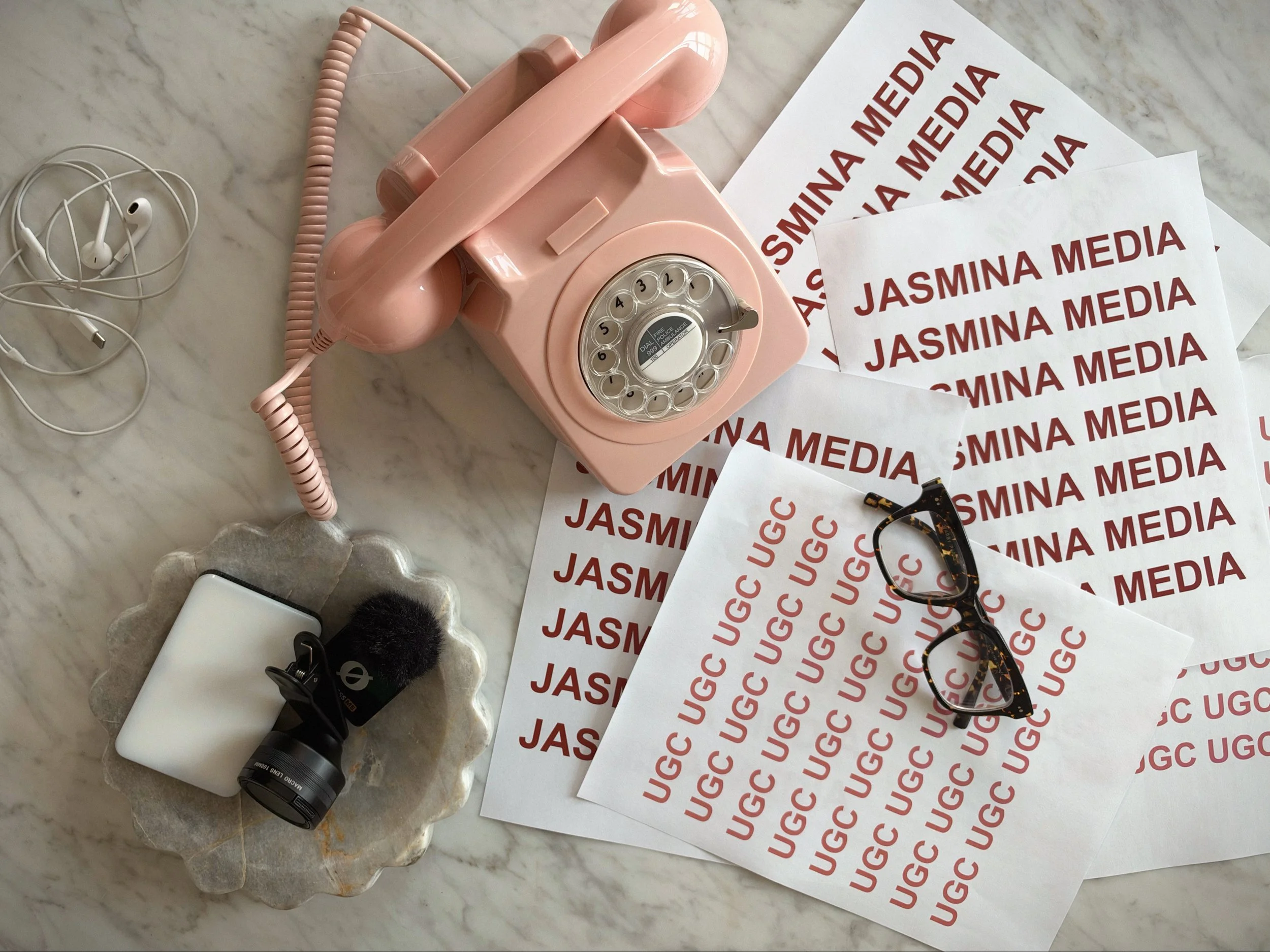 A pink vintage rotary telephone, a pair of glasses with a tortoise shell frame, several sheets of paper with red text reading 'JASMINA MEDIA,' a white power adapter, black earbuds, and a small furry black object on a marble surface.