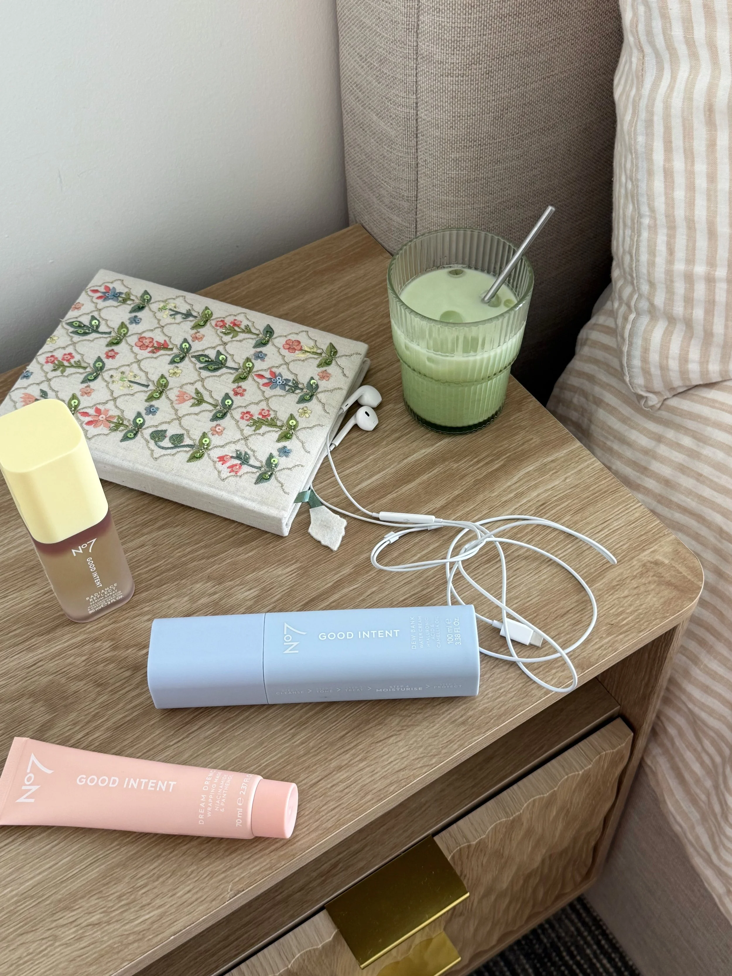 A wooden bedside table with various skincare products, a glass of green liquid with a metal straw, a cloth with floral embroidery, and tangled white earphones.