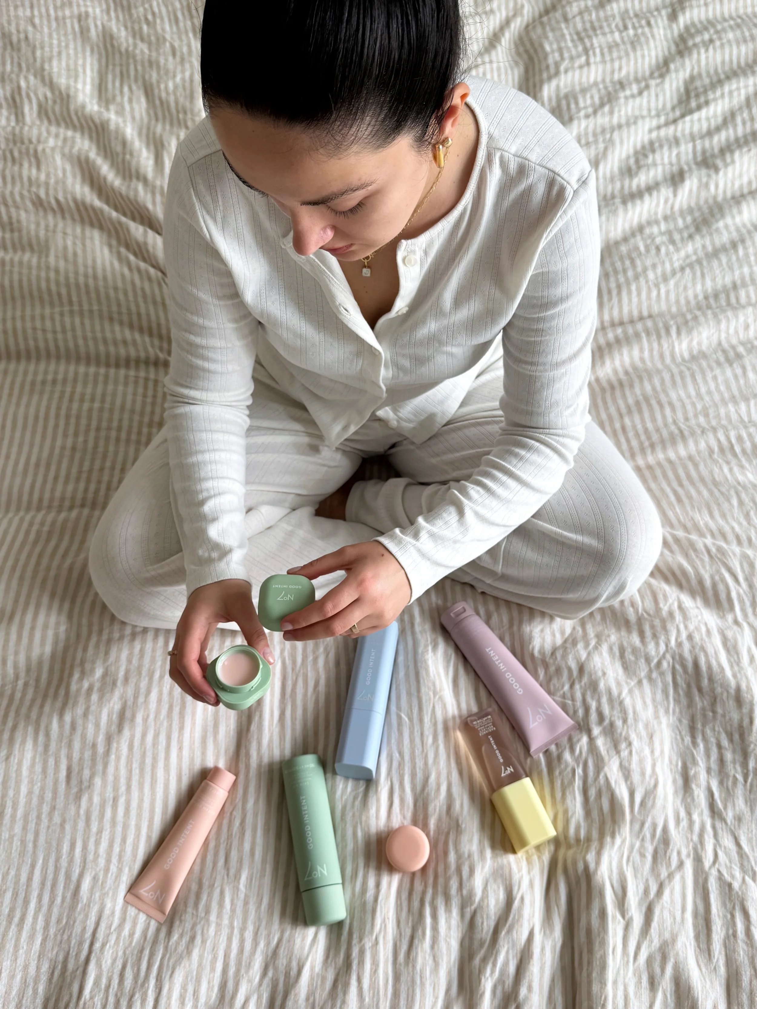 A woman sitting on a bed in white pajamas, surrounded by pastel-colored skincare or cosmetic bottles, some of which she is opening or touching.