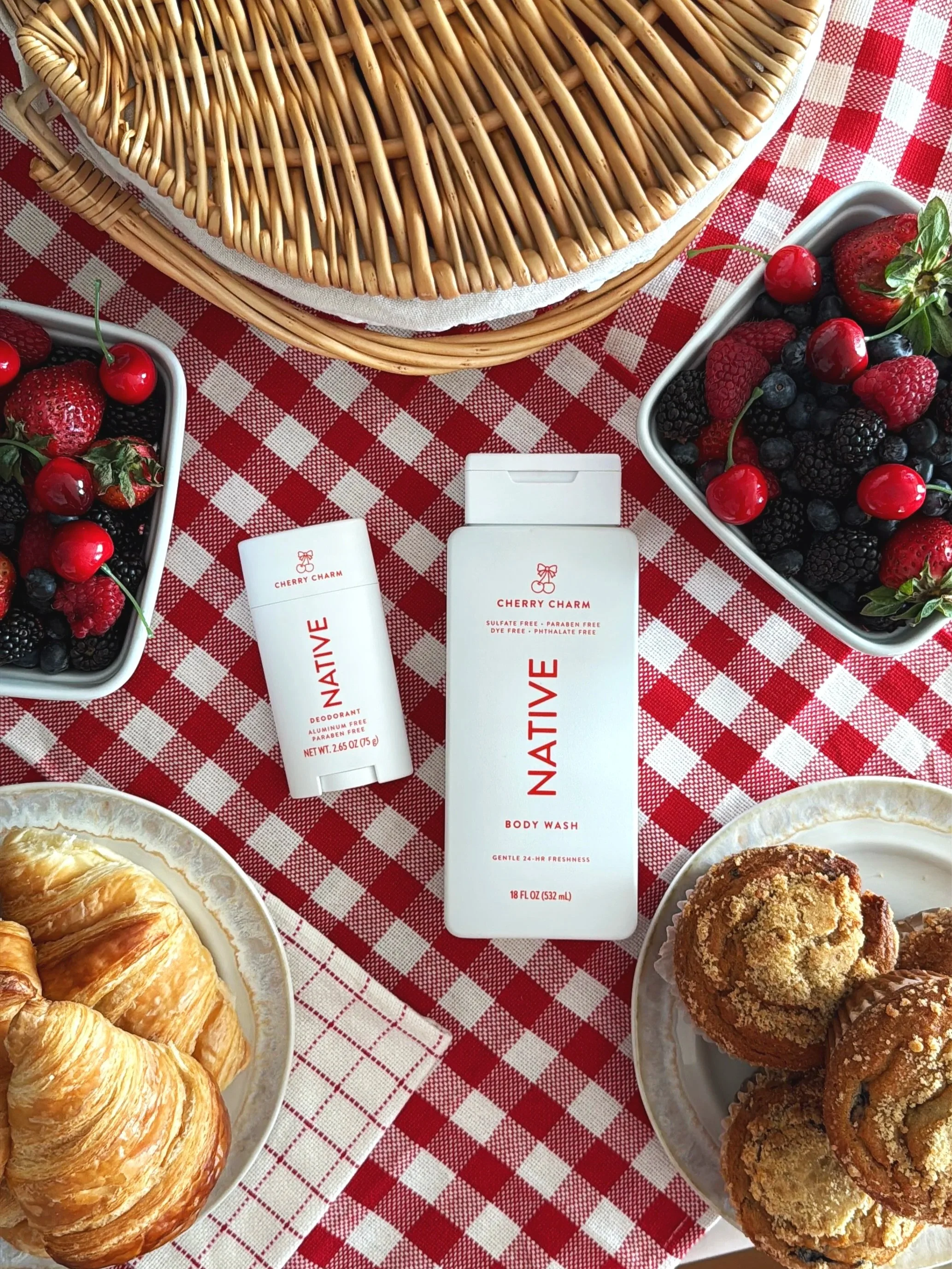 A table with a red and white checkered tablecloth featuring plates of croissants, a bowl of mixed berries including strawberries, blueberries, raspberries, and cherries, along with skincare products labeled "Cherry Charm" body wash and deodorant.