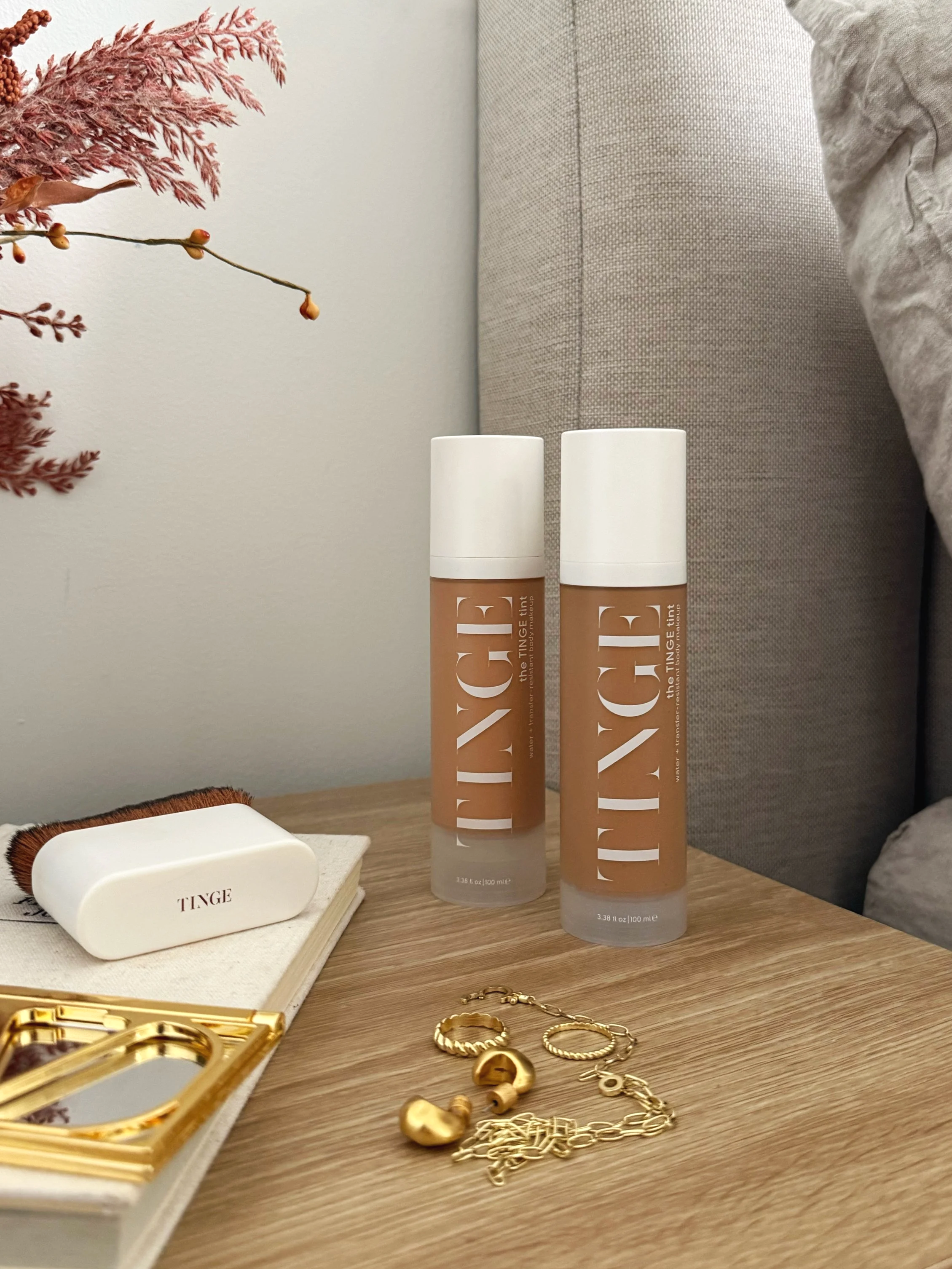 A wooden table holding two bottles of tanning serum, a small white soap with 'Tinge' printed on it, gold jewelry including earrings, rings, and chains, a gold makeup mirror, and a notebook with a brush on top. In the background, there is a grey fabri