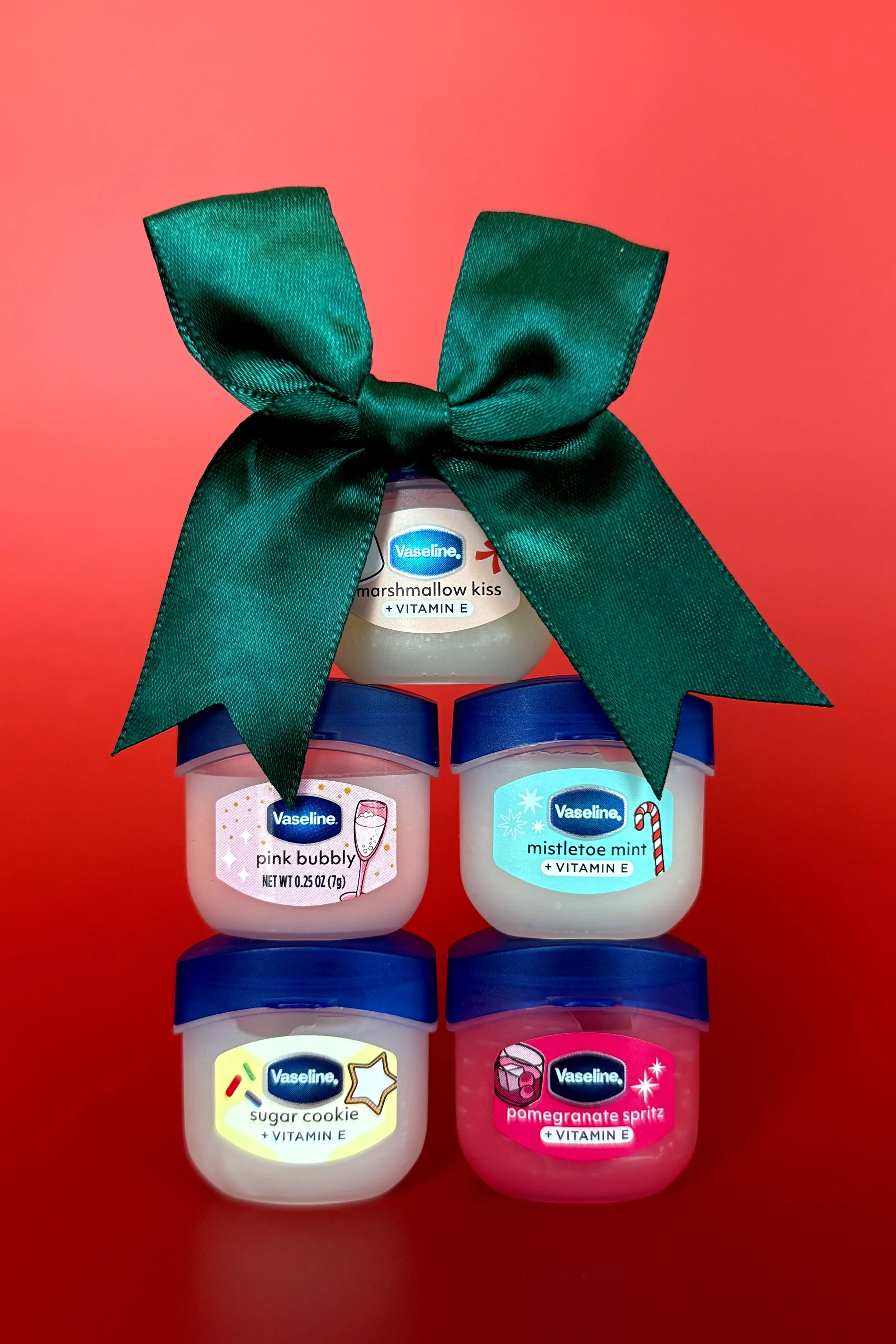 A group of five Vaseline lip balm containers arranged against a red background. Each container has a different flavor or variant, including marshmallow kiss, pink bubbly, mistletoe mint, sugar cookie, and pomegranate spritz, with colorful labels and 