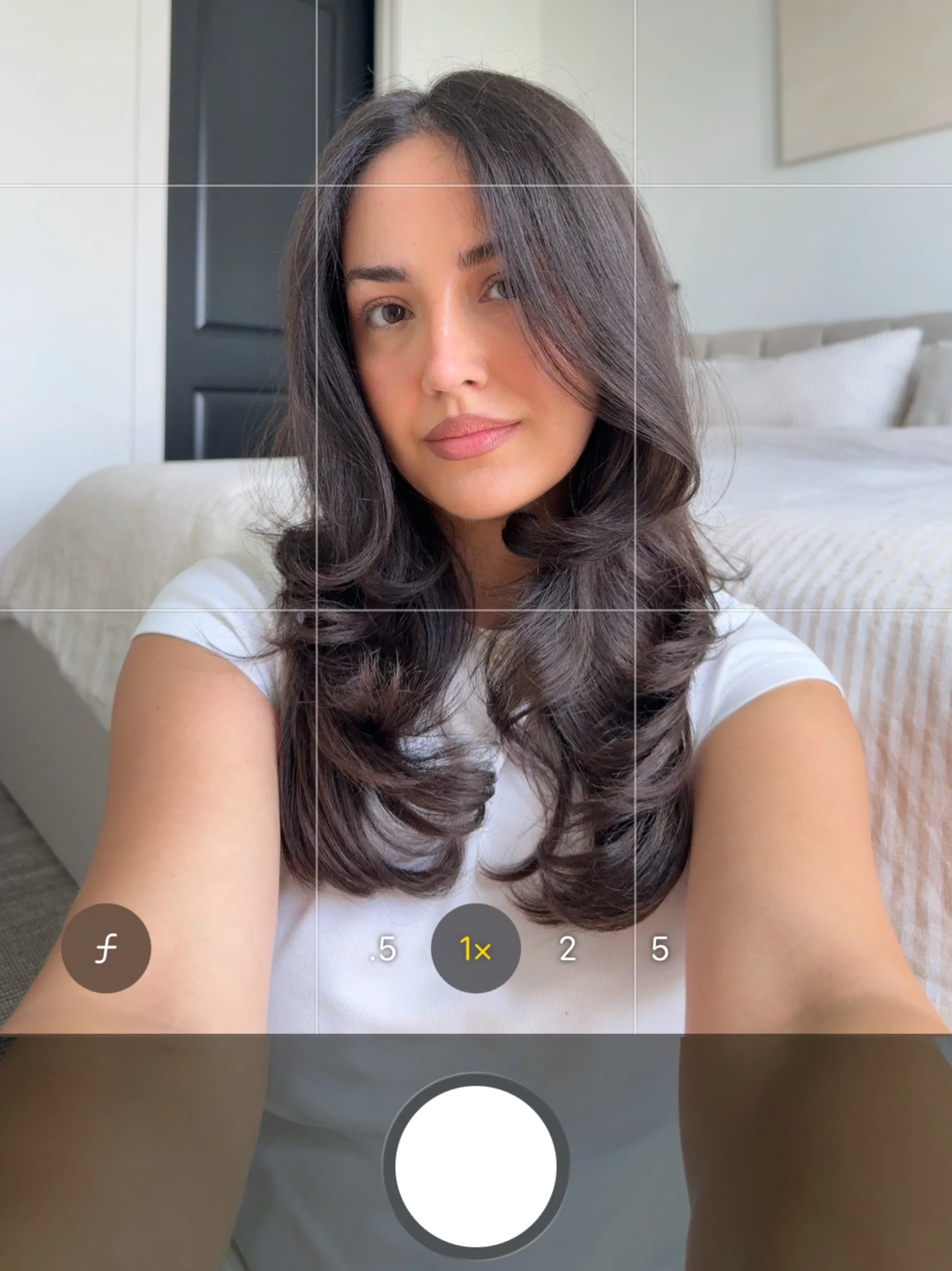 A young woman with long dark hair and light skin taking a selfie in a bedroom with a bed and pillows in the background.