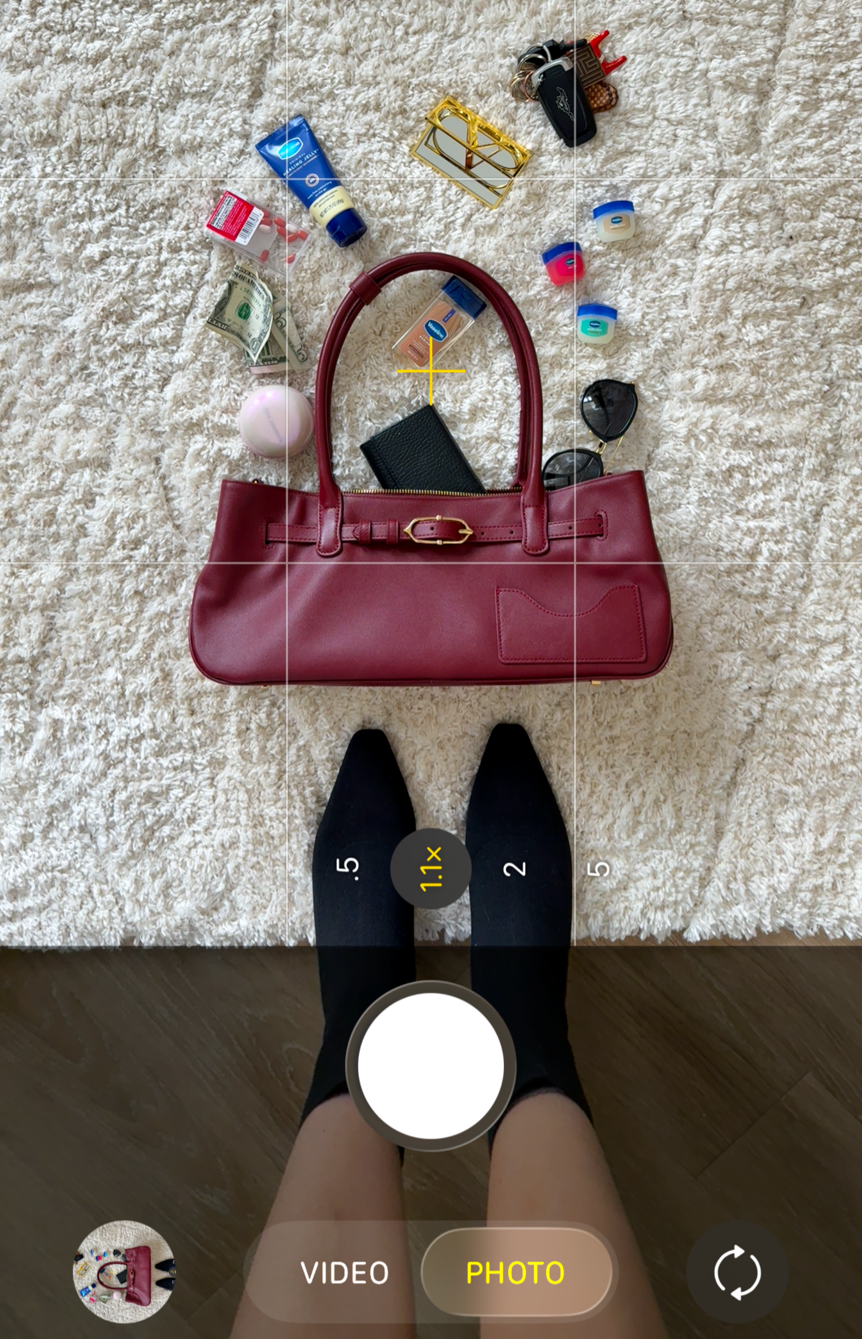 Top-down photo of a person standing on a hardwood floor, with a red handbag on the floor, along with various cosmetic and personal items, a pair of sunglasses, and a wallet. The person is taking a selfie with a smartphone camera app.