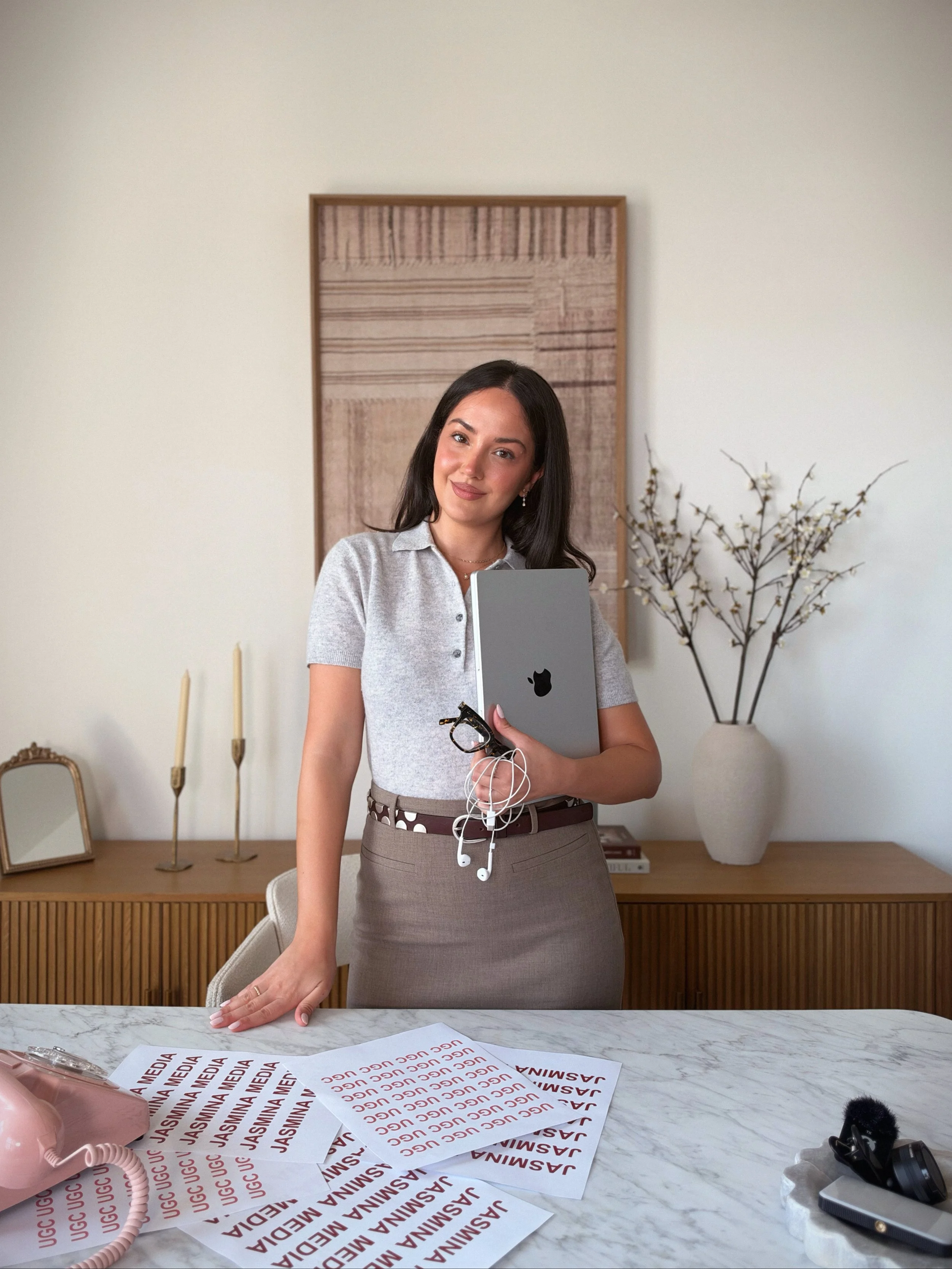A woman standing behind a white marble table holding a laptop and some earphones, with printed sheets of paper on the table. The background features a wooden sideboard, decorative candlesticks, framed artwork, and a large vase with branches.