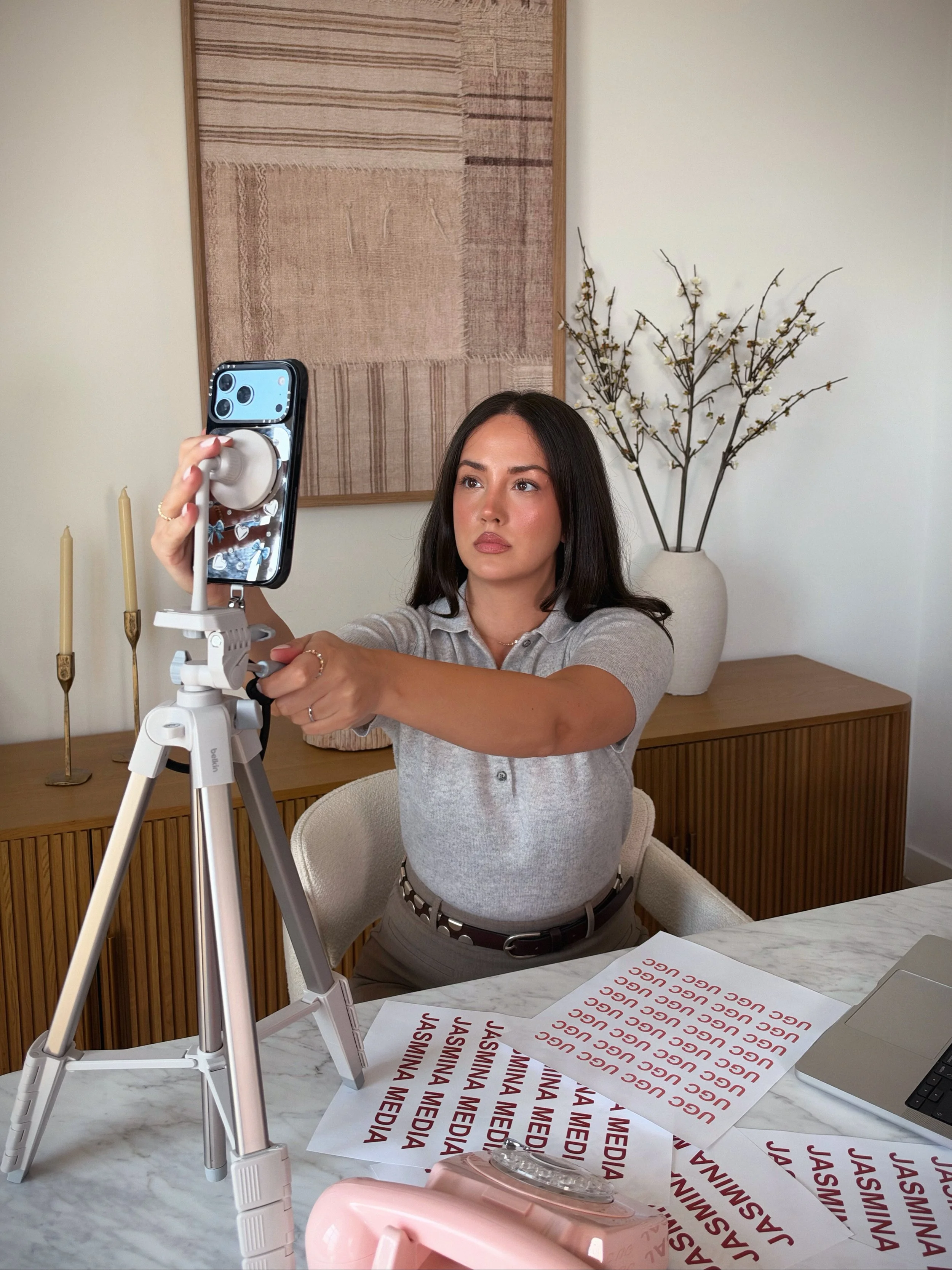 A woman with dark hair and a light gray shirt sitting at a white marble table, taking a selfie with her smartphone mounted on a tripod. On the table are sheets of paper with the text 'JASMINA MEDIA' repeatedly printed in red. In the background, there is a wooden sideboard with two candles, a large beige vase with branches and small white flowers, and a woven wall hanging.