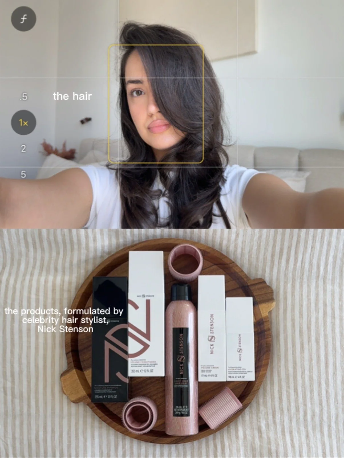 A woman taking a selfie with a messy hairstyle in a bright room with a beige sofa in the background. On a wooden tray below, there are various hair styling products in pink and black packaging, some boxes, a pink hair roller, and a pink hairbrush.