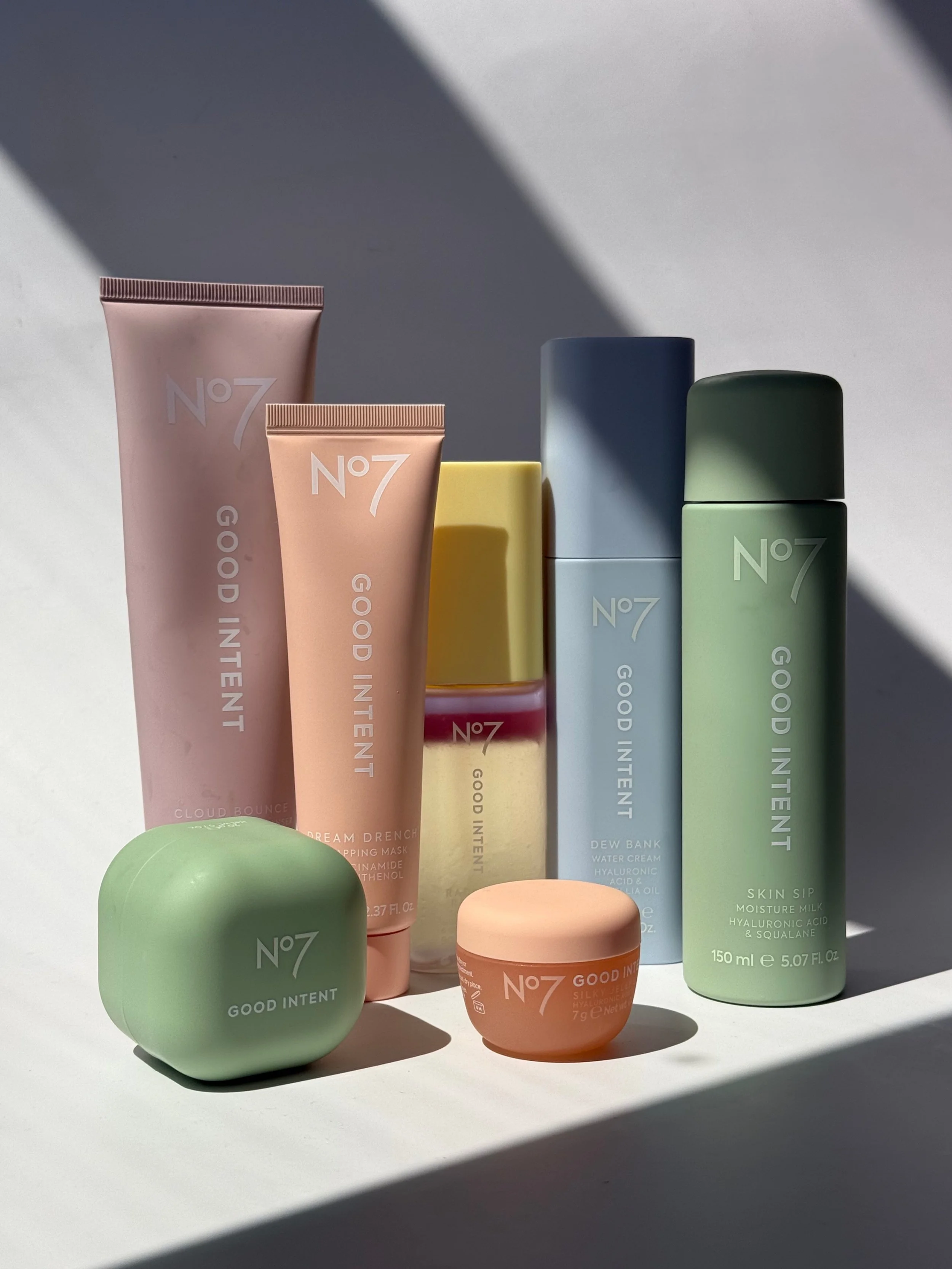 Collection of skincare products from No7, including tubes, bottles, and jars in pastel colors like pink, yellow, blue, green, and peach, arranged on a white surface with shadows cast across the items.