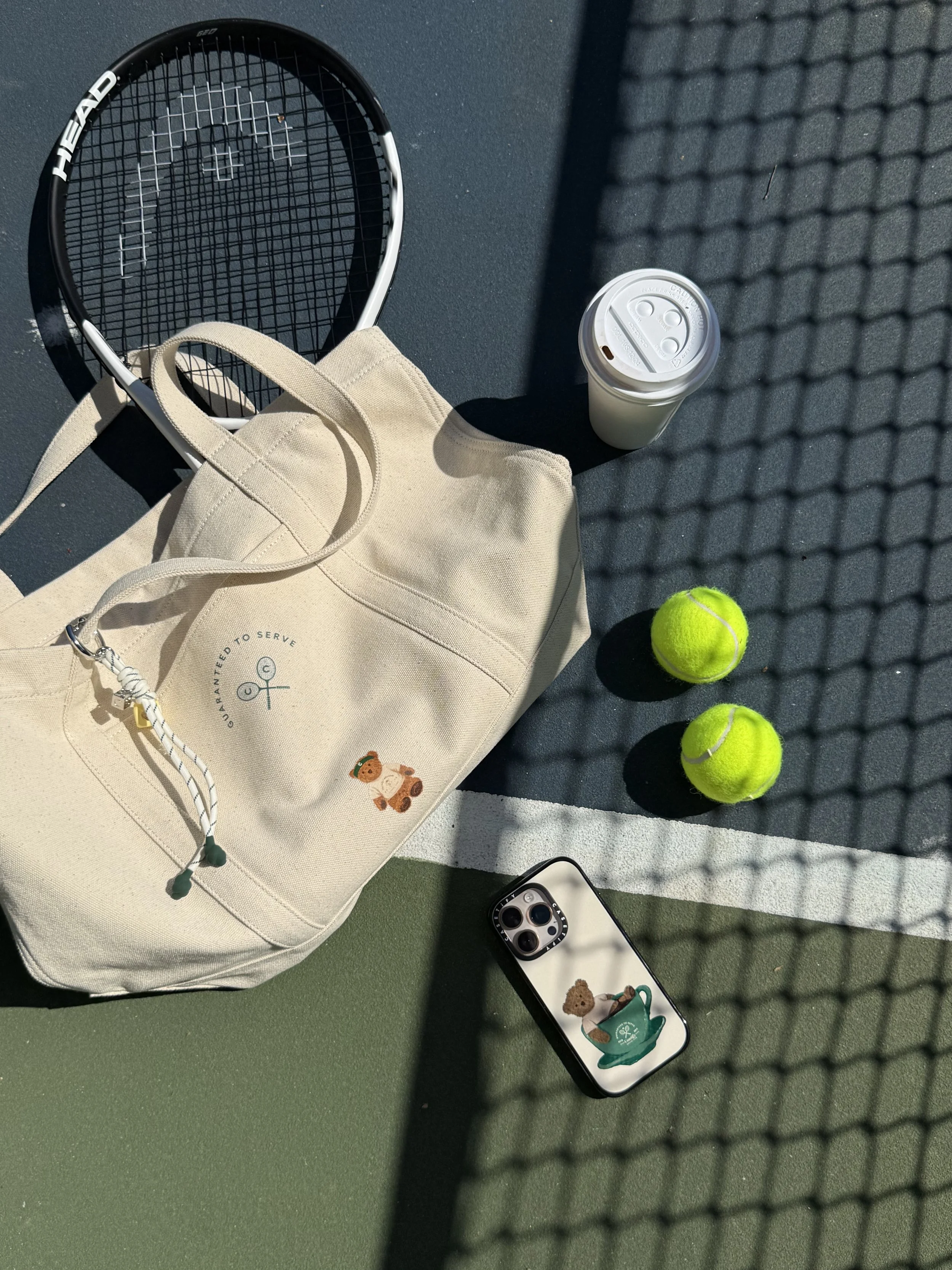 A tennis court with tennis balls, a tennis racket, a beige tote bag with a teddy bear illustration, a disposable coffee cup, and a smartphone displaying a teddy bear illustration on its screen.