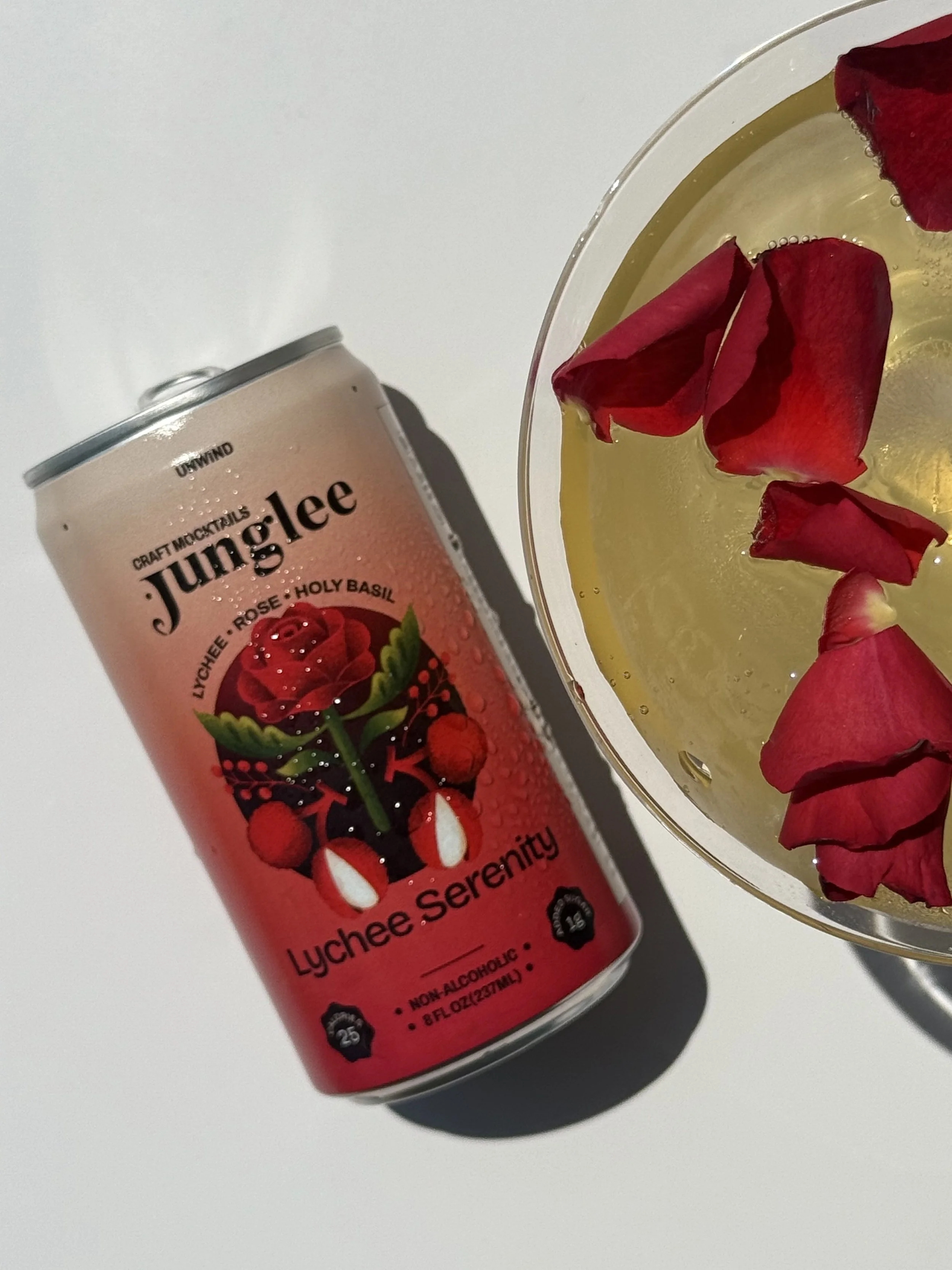 A can of craft cocktail sparkling beverage called Lychee Serenity with a pink label featuring lychee fruit images and roses, and a glass with yellow liquid and red rose petals.