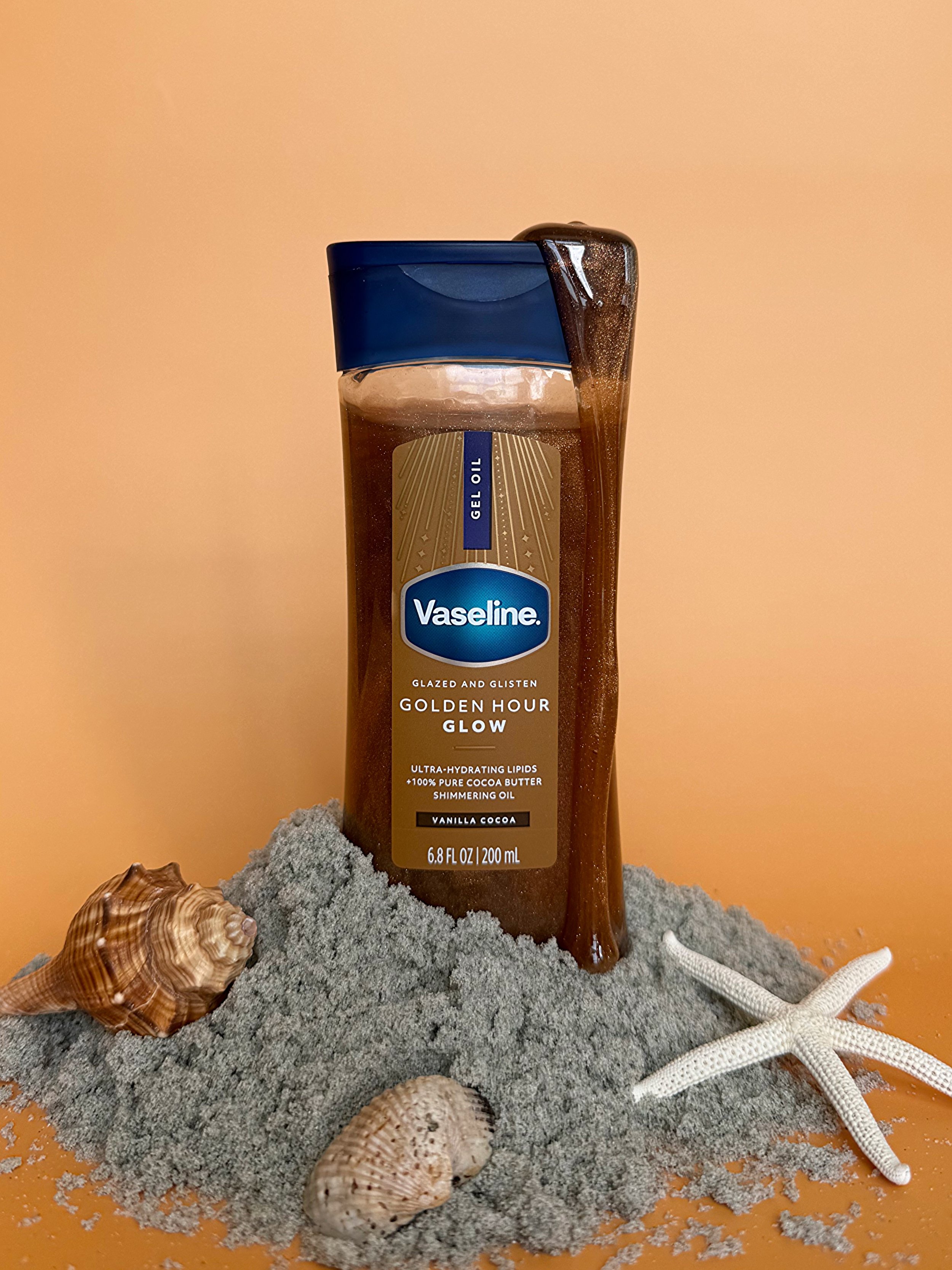 A bottle of Vaseline Golden Hour Glow gel oil placed on sand with seashells and starfish, against an orange background.