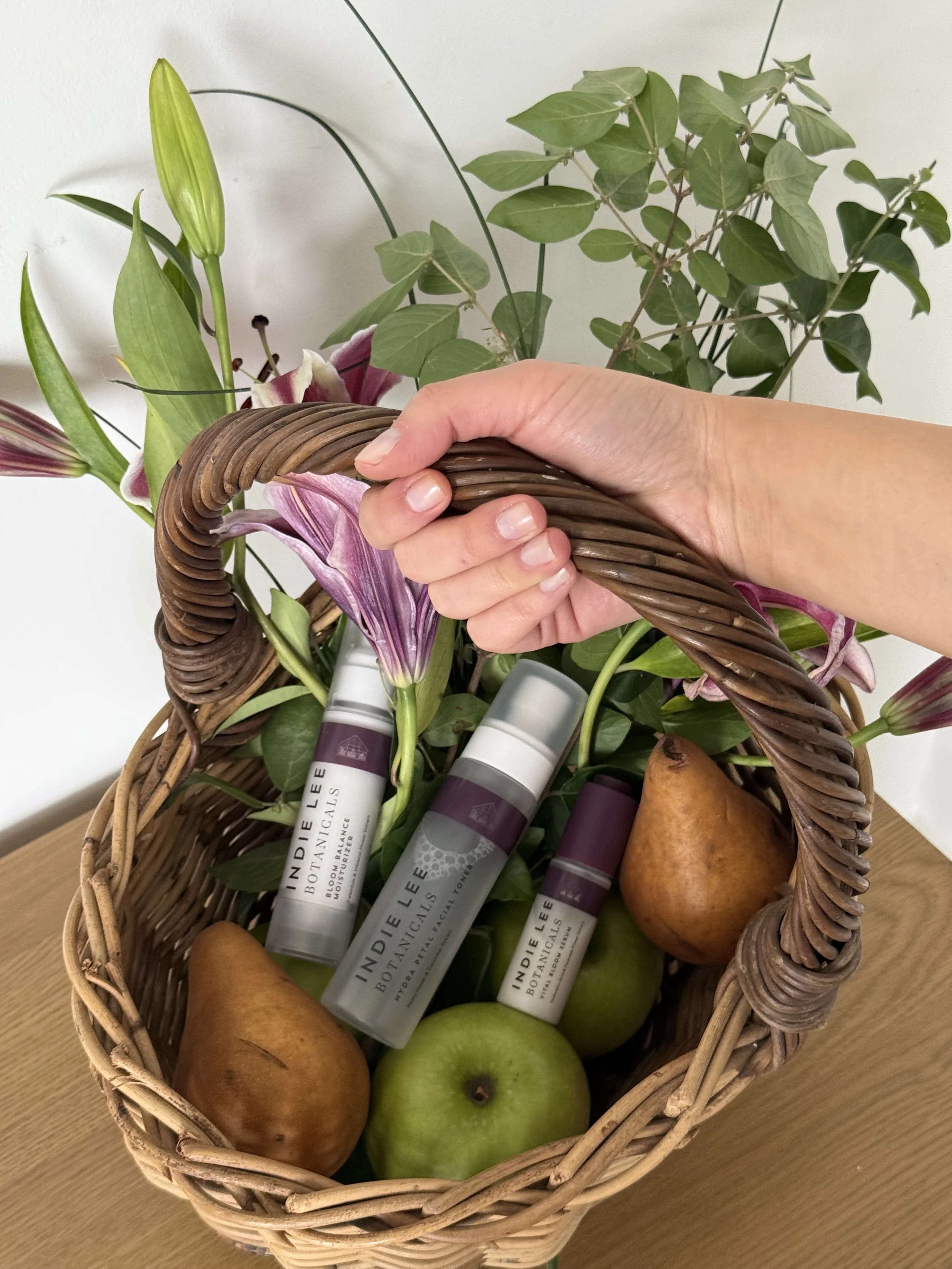 A wicker basket contains green apples, pears, and several tubes of skincare products with white labels, on a wooden surface.
