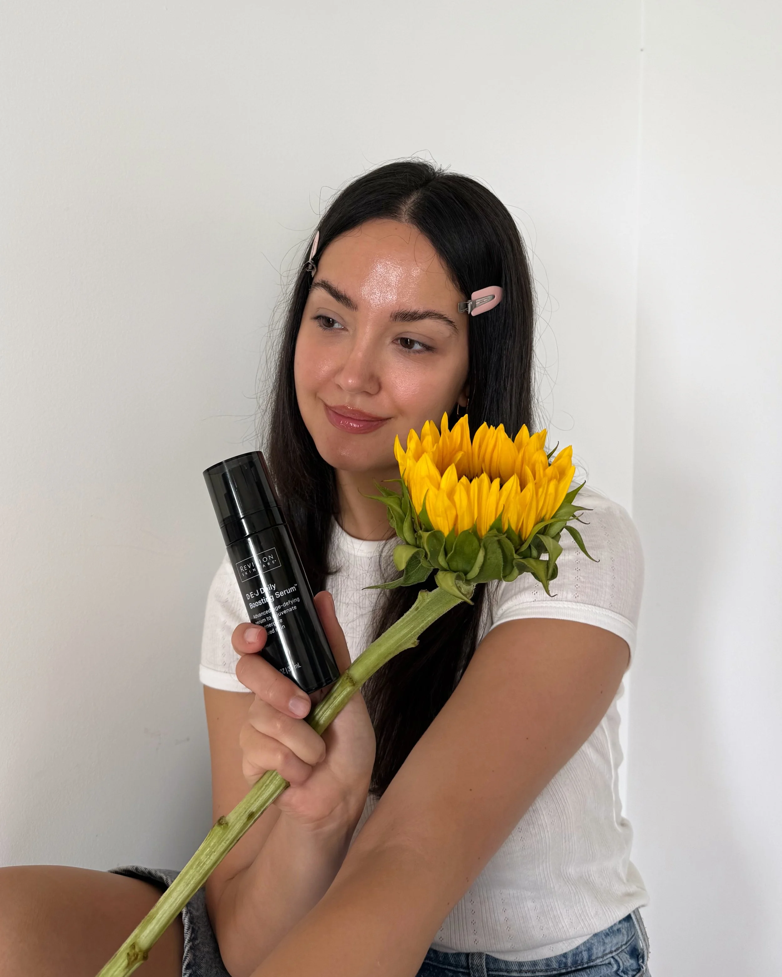 A woman with long dark hair, wearing a white t-shirt and denim shorts, holding a sunflower, a black bottle, and a skincare serum against a plain white wall.