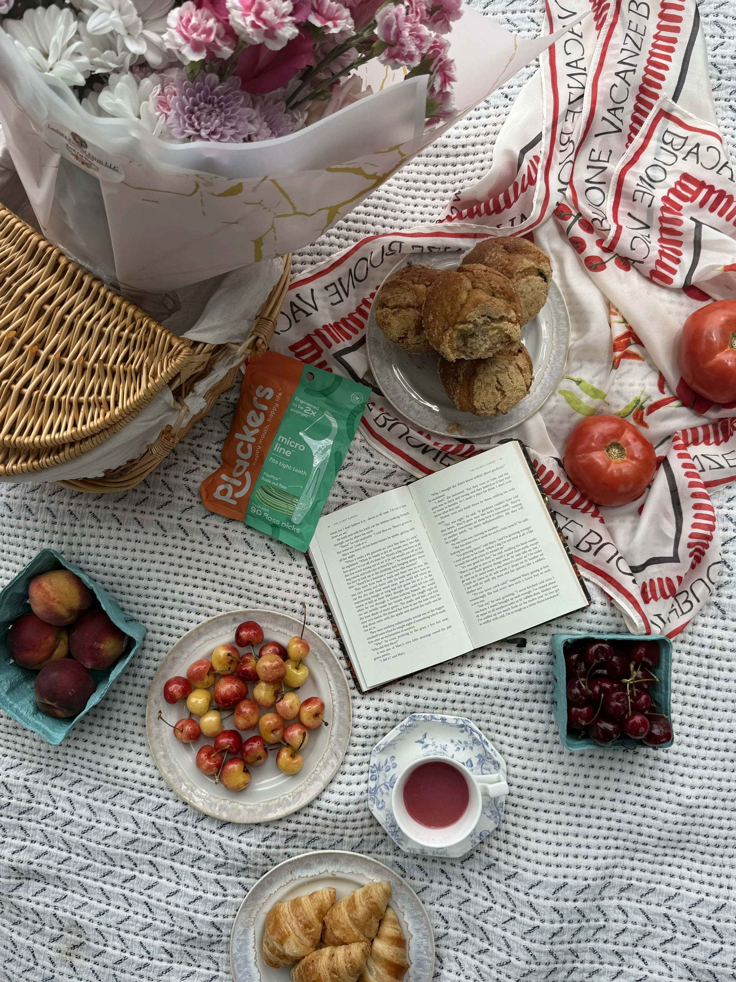 A picnic spread on a checkered fabric with flowers, pastries, fruits, and a cup of tea.