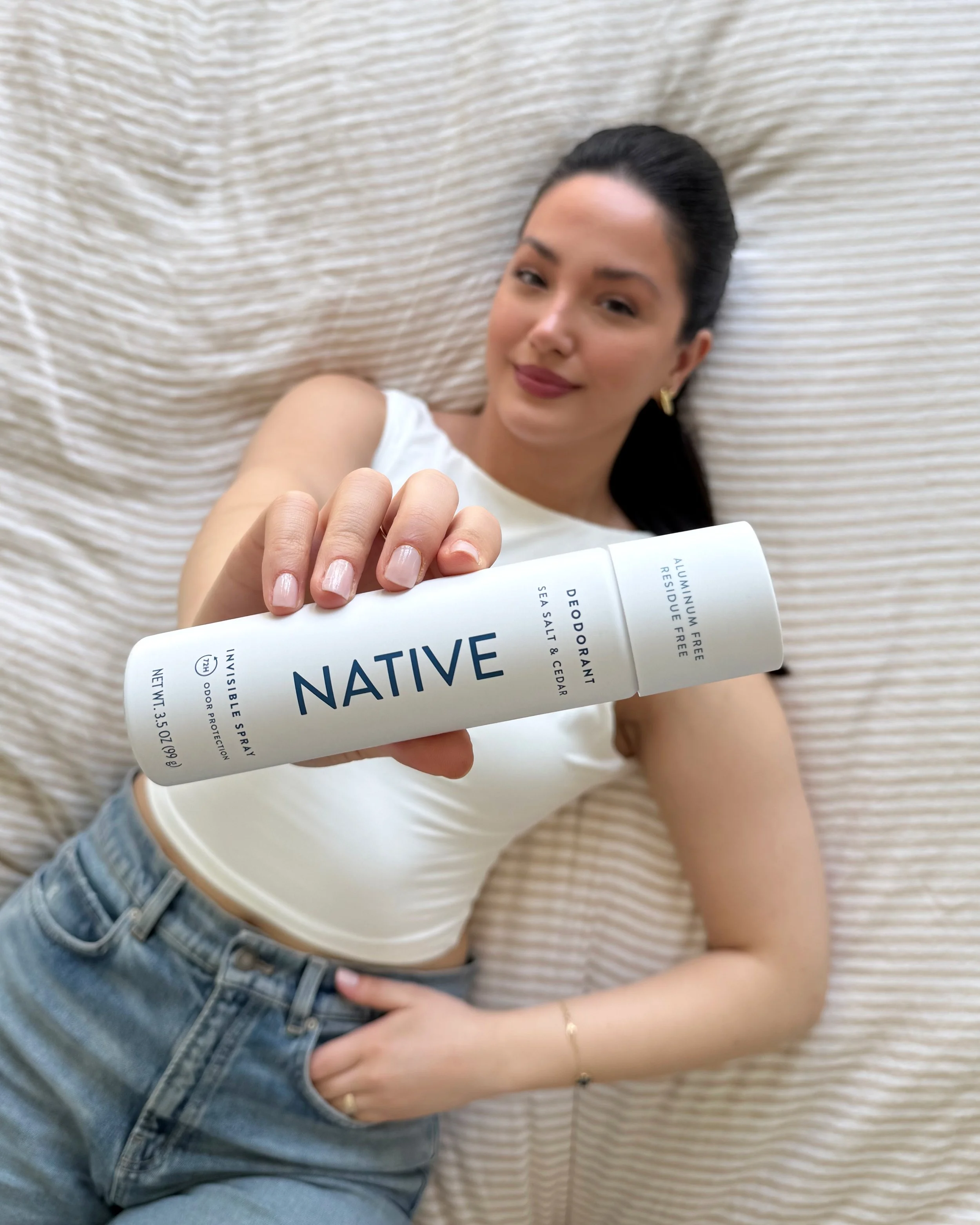 Young woman lying on a bed with beige textured sheets, holding a white bottle of NATIVE deodorant. She is wearing a white sleeveless top and blue jeans, with dark hair pulled back and gold earrings, smiling softly at the camera.