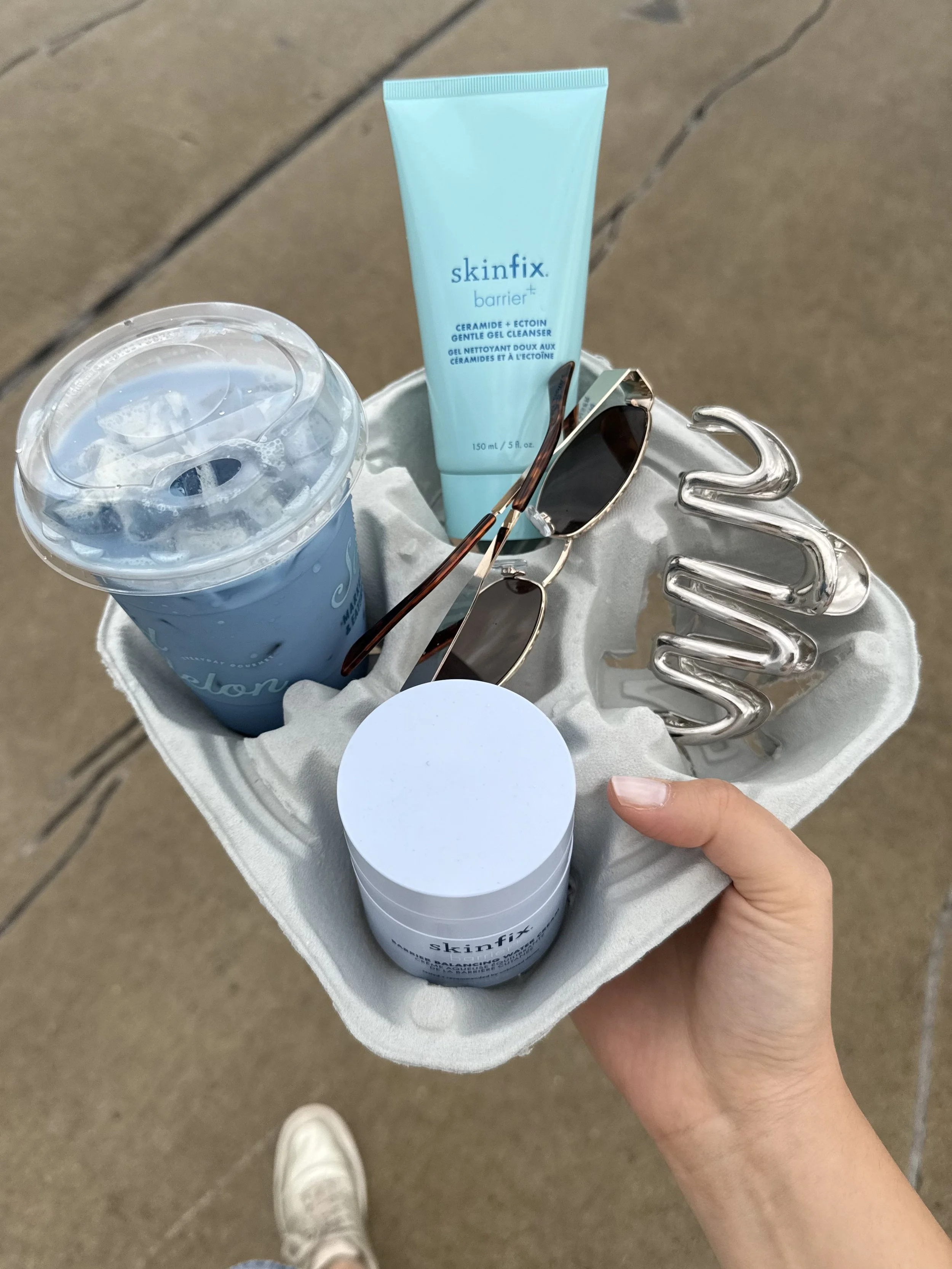 A hand holding a white container with skincare products, including a blue tube of Skinfix barrier+ gel cleanser, a cup of ice water in a blue cup, and a pair of sunglasses, with a metallic S-shaped sculpture in the background.