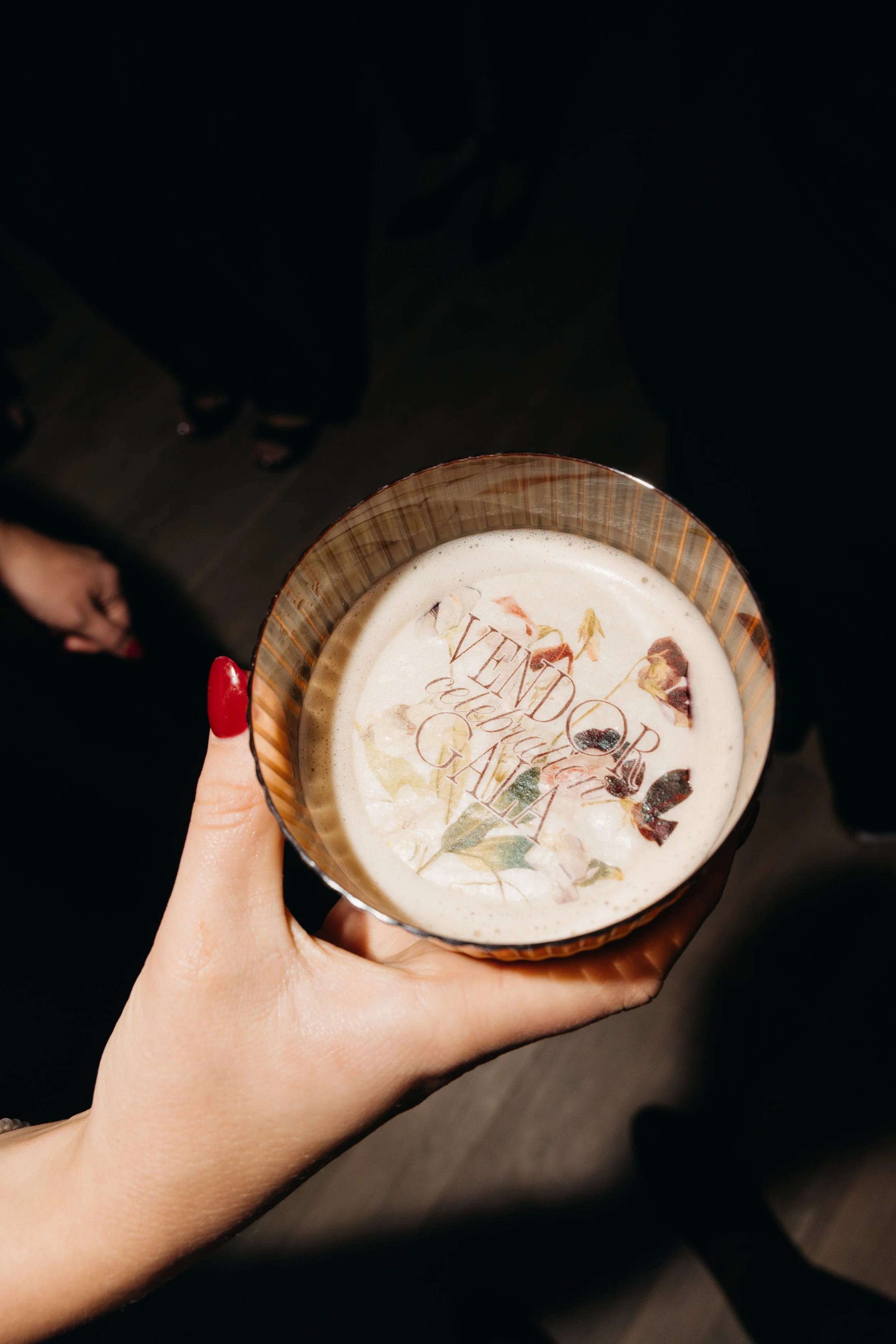Hand holding a vintage-style beverage cup with a label that reads names and ingredients, against a dark background, with people and shoes visible in the background.