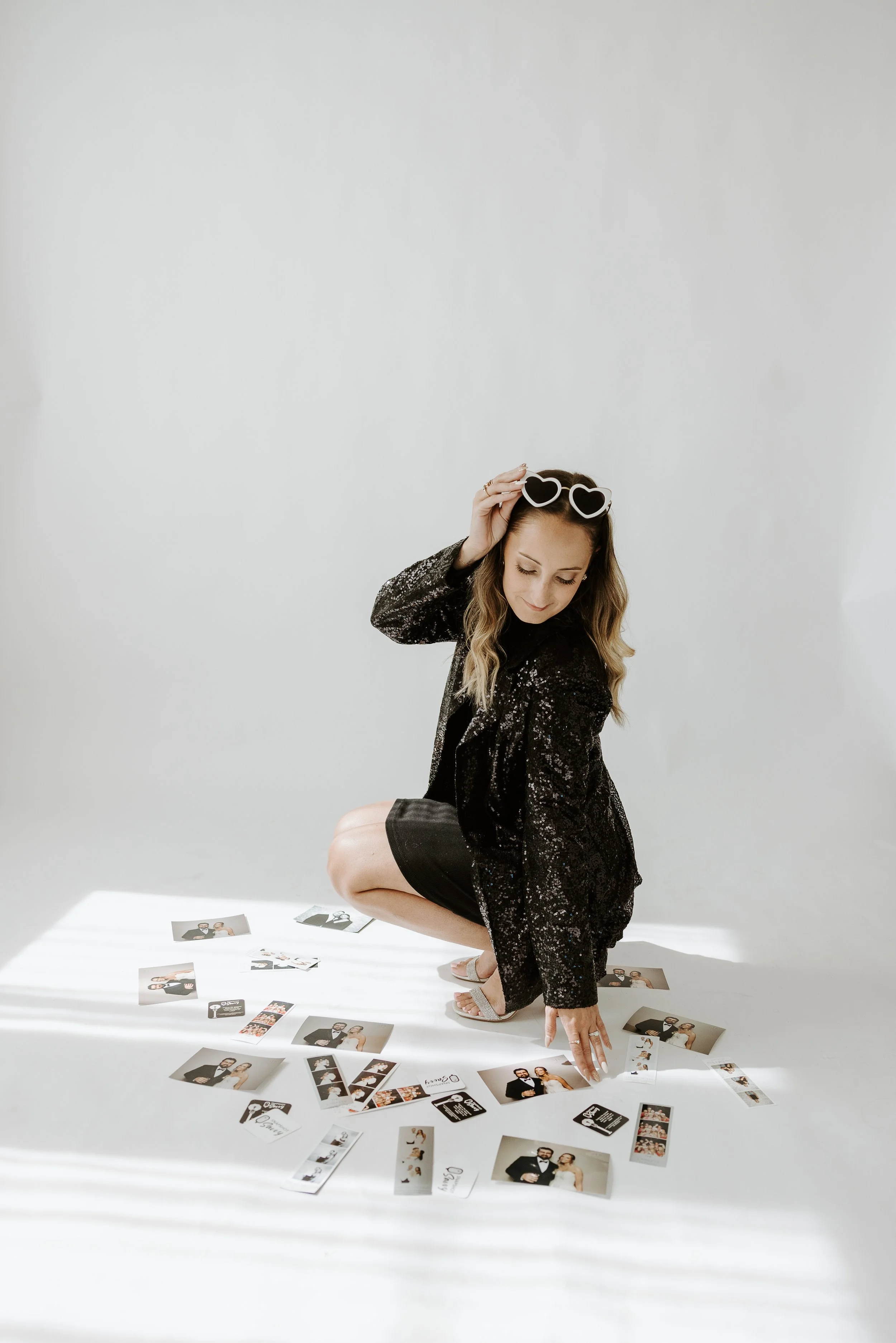 A woman in a black sequined dress and heart-shaped sunglasses is crouching, pointing with one hand, surrounded by wedding photos and cards on a white floor against a plain white wall.