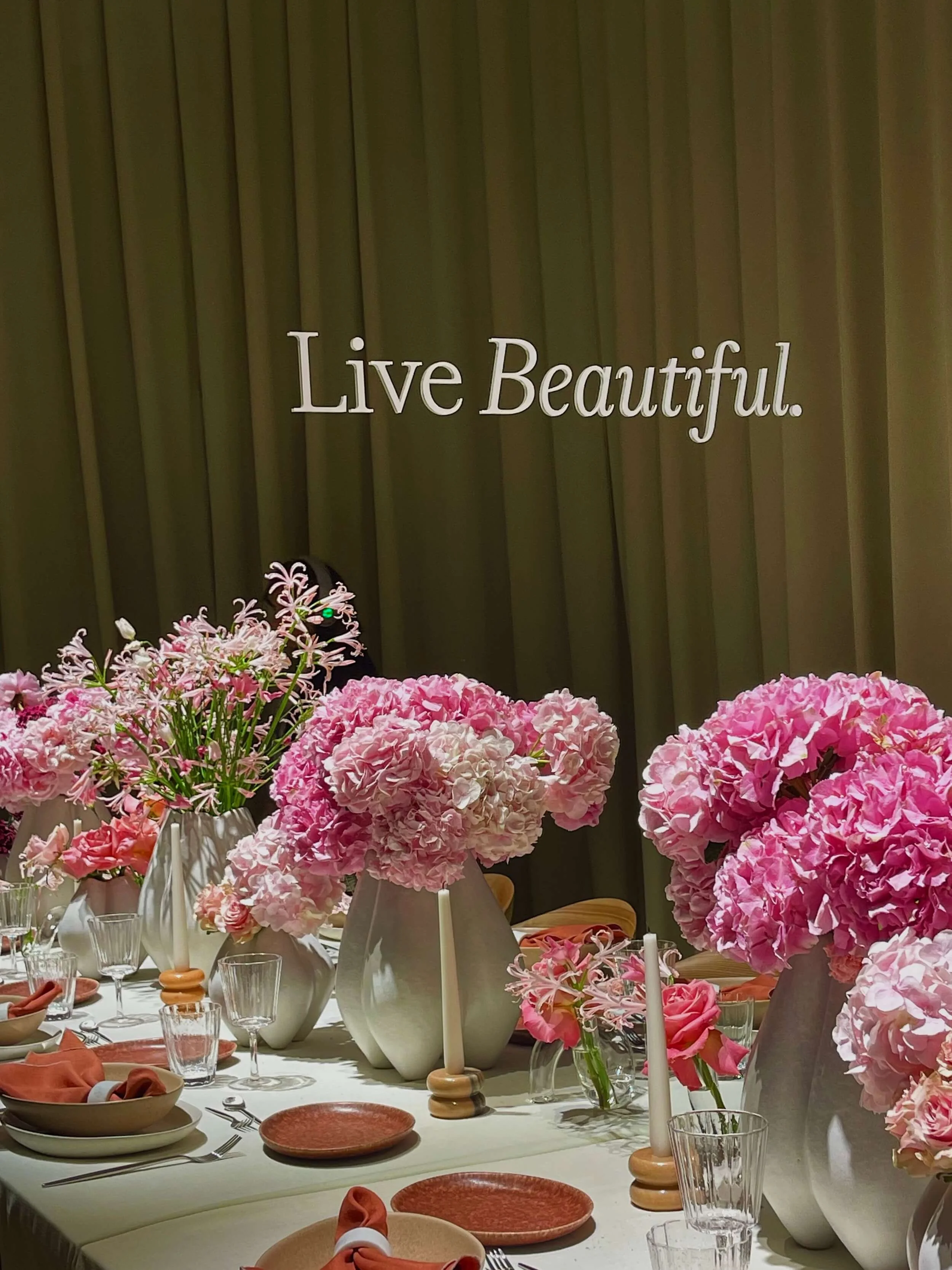 Decorated dining table with pink flowers in vases, candles, glassware, and orange napkins, set for a meal with a 'Live Beautiful' sign on a green wall in the background.