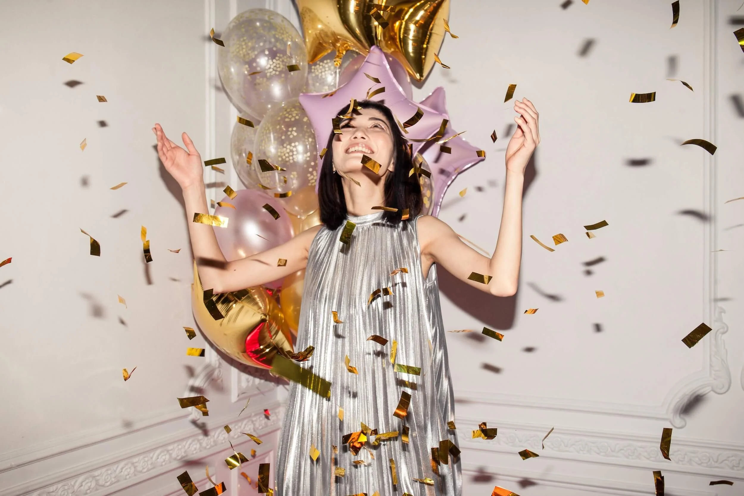 A woman celebrating with gold and pink balloons and confetti falling around her against a plain wall.