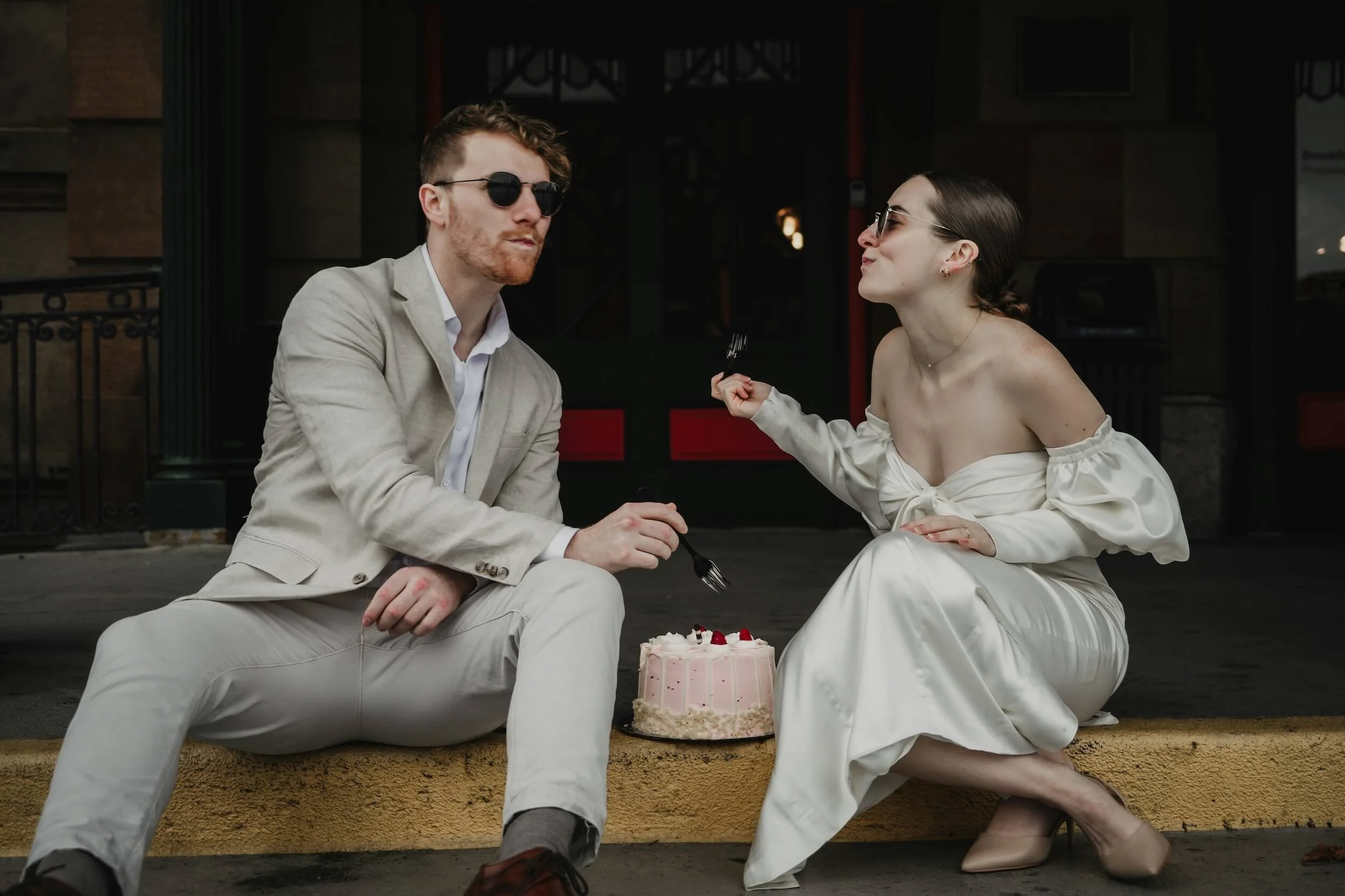 A couple sitting on the sidewalk, dressed elegantly, with a cake in front of them, in a moment that appears romantic.