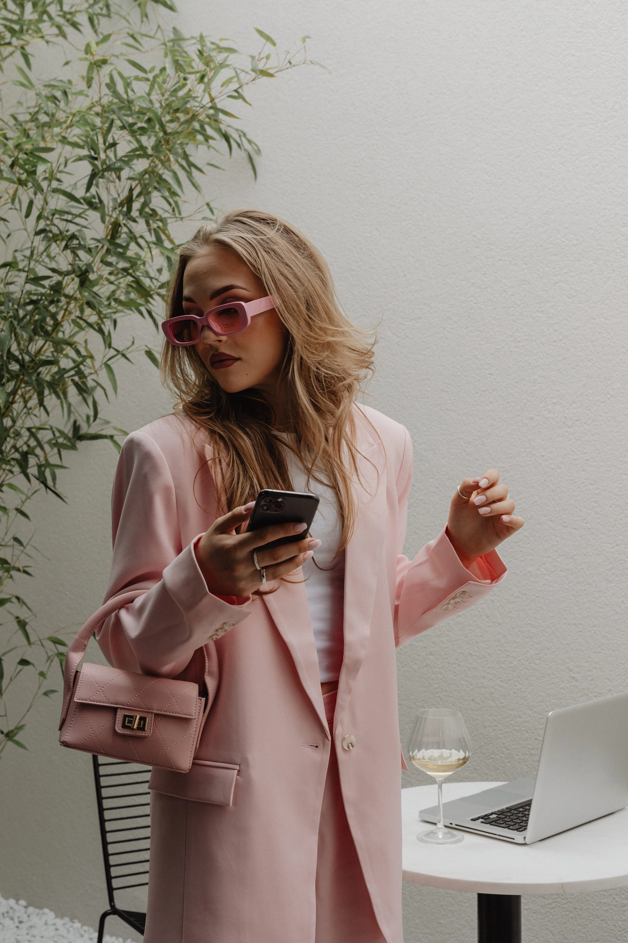 A woman in a pink suit with pink sunglasses, holding a smartphone, standing near a table with a glass of white wine, a laptop, and a chair in a minimalist setting with a potted plant.