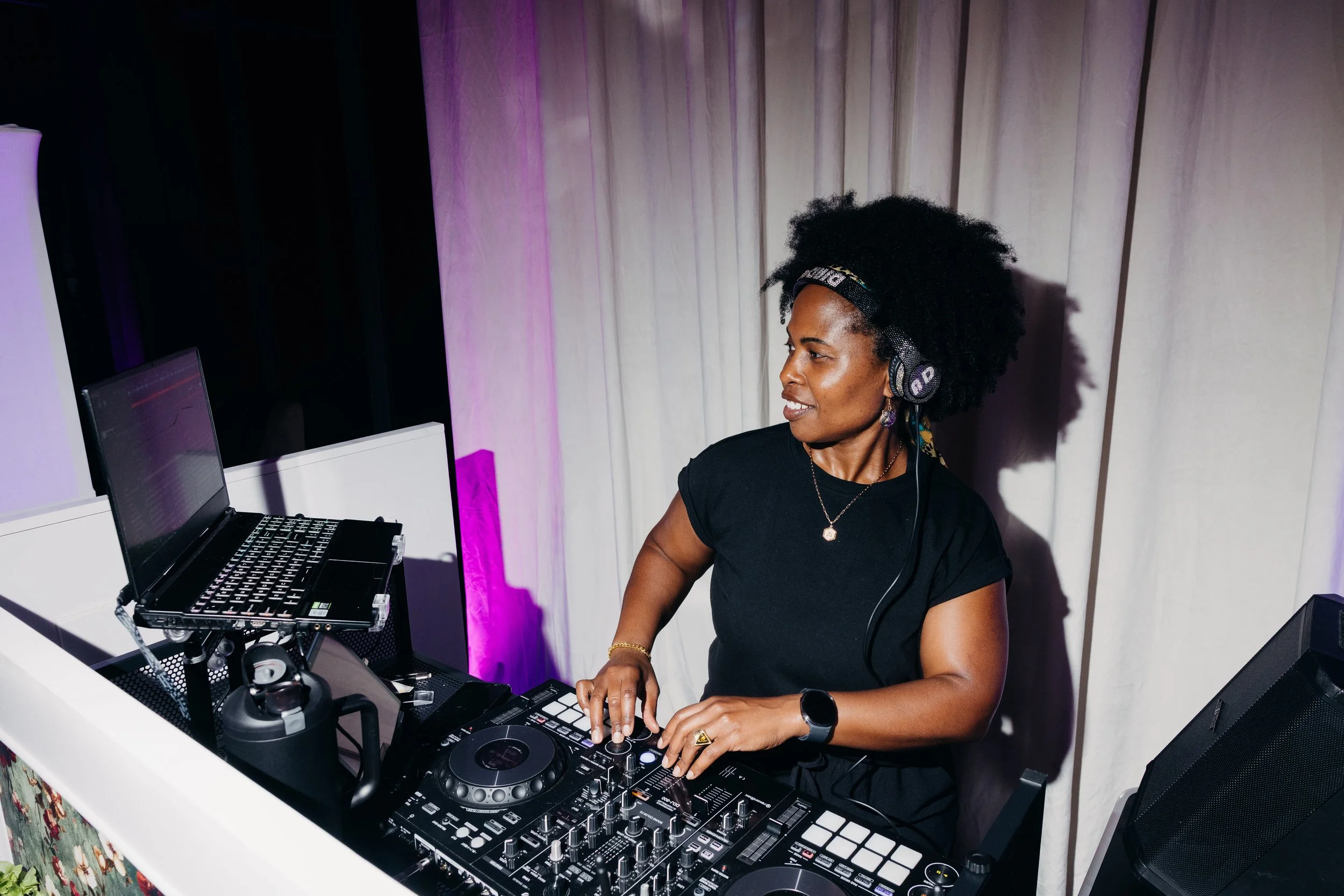 A woman with natural curly hair wearing headphones and a black t-shirt, DJing at a table with DJ equipment, in a venue with white curtains and purple lighting.