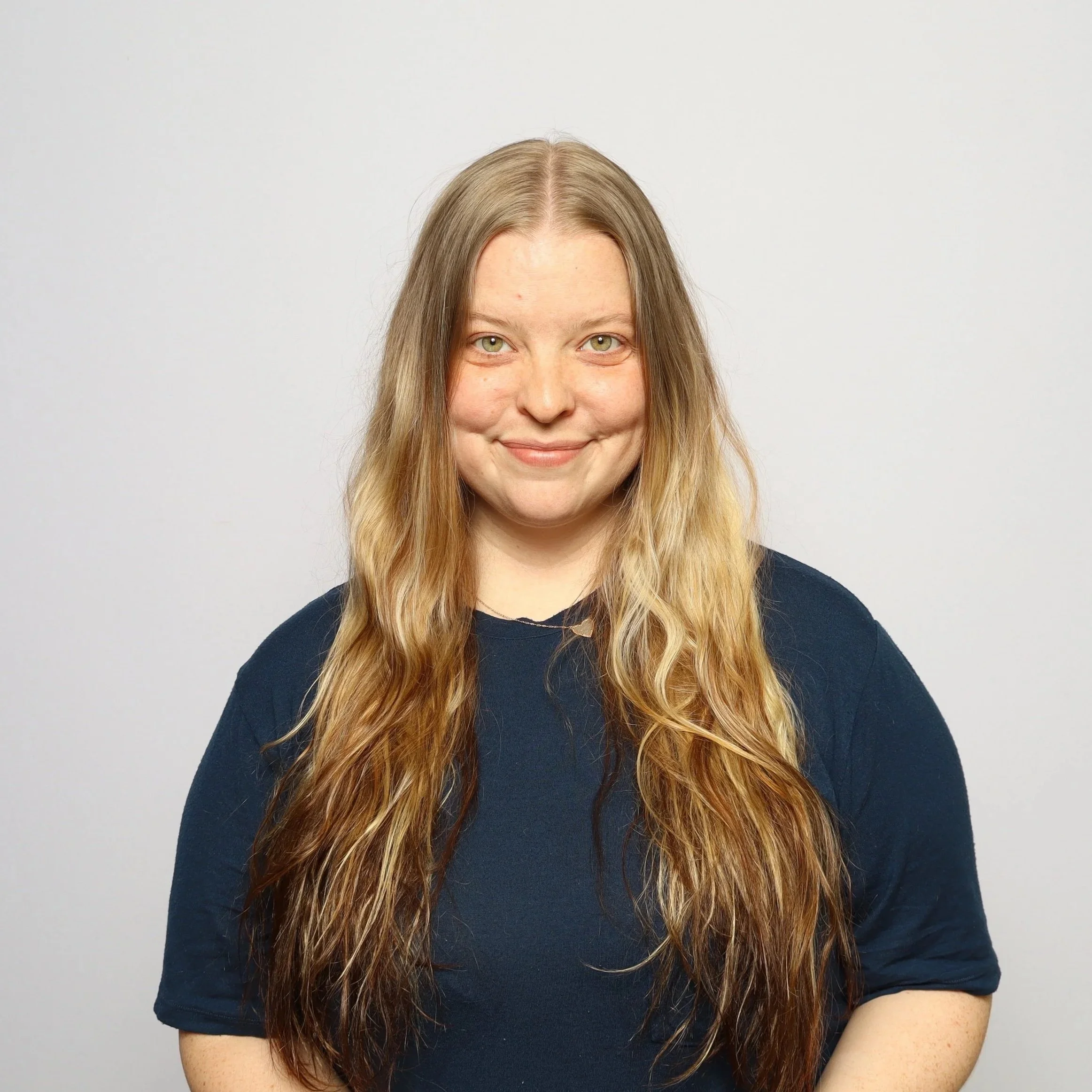 A young woman with long, wavy blonde hair and light green eyes smiling against a plain white background.