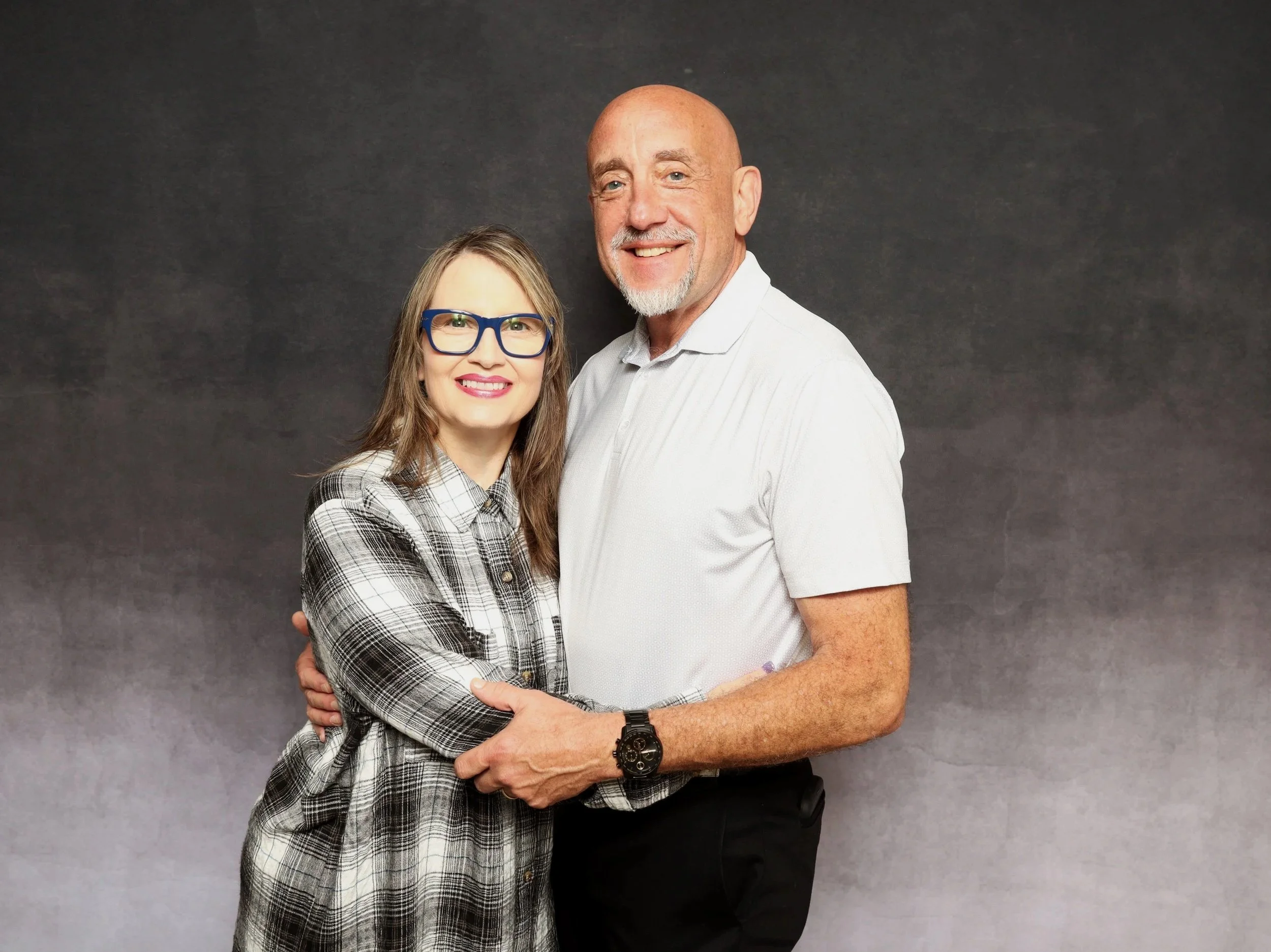 A man and woman are standing close together and smiling at the camera against a dark gray background. The woman has long brown hair, is wearing blue glasses, and a black and white plaid shirt. The man is bald, with a short white beard, and wearing a white polo shirt.