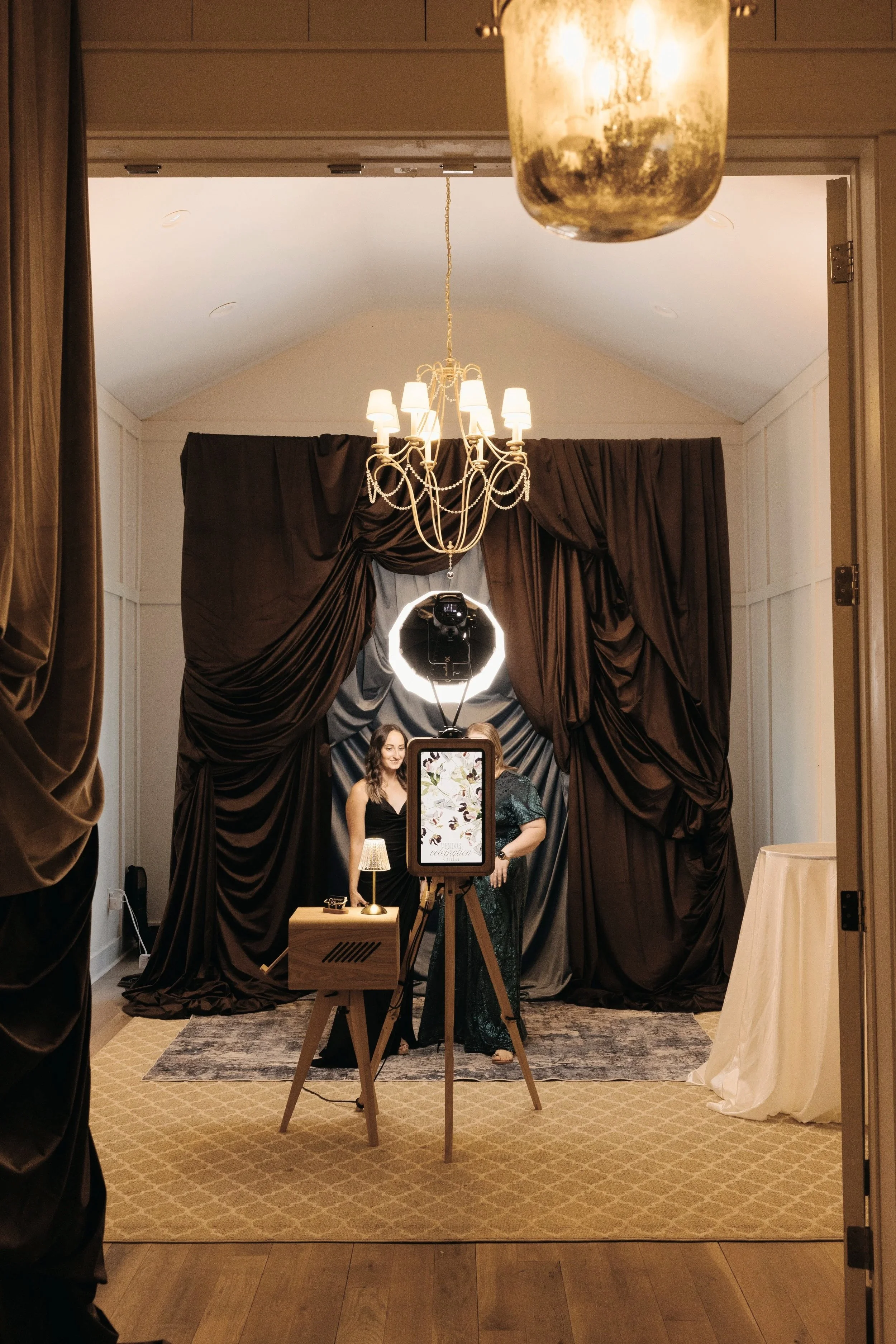 People taking a photo of a woman in formal attire at a photo booth with elegant draped curtains, a chandelier, a ring light, and a calender on a stand.