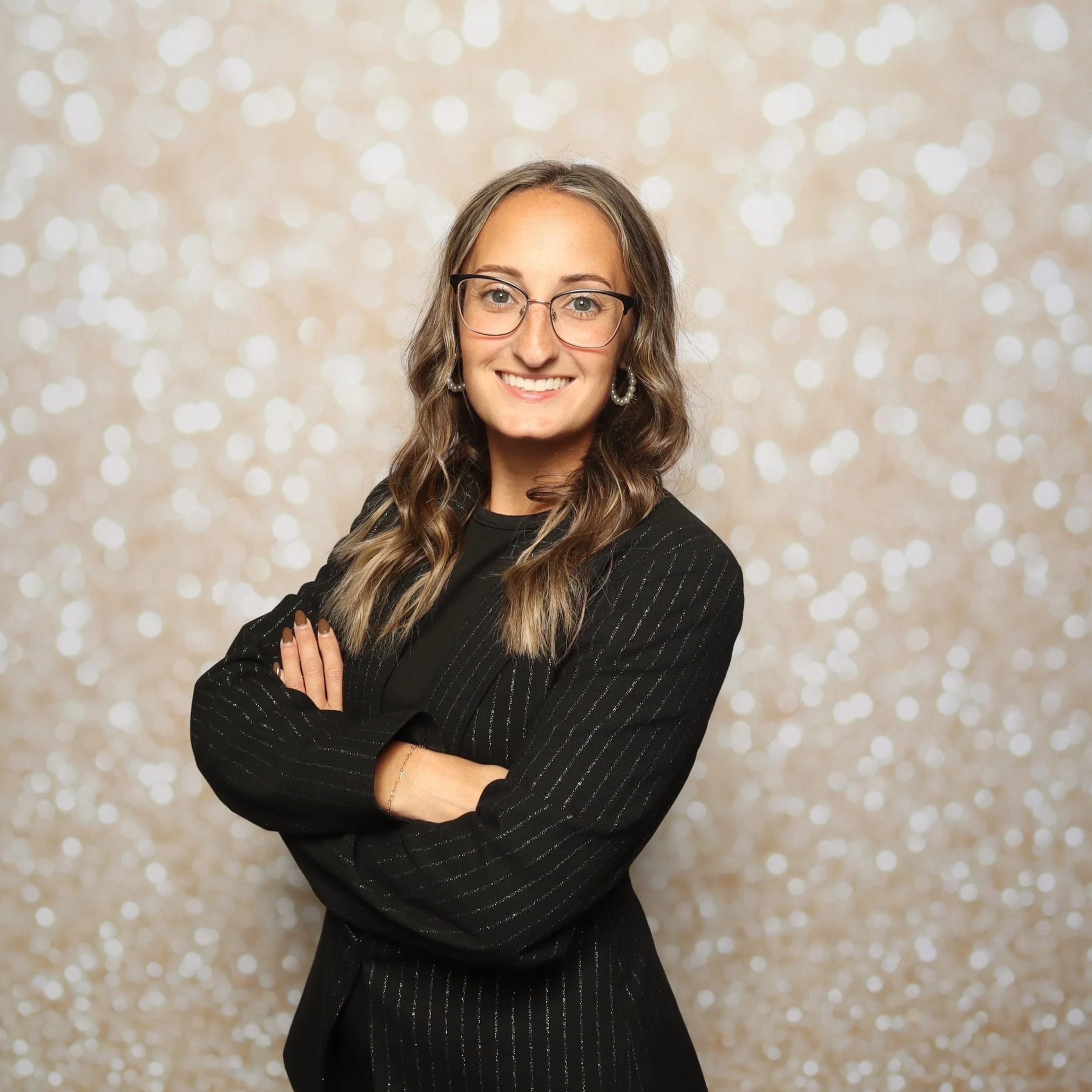 A woman with long wavy hair, glasses, and earrings, smiling, wearing a black pinstripe blazer, standing in front of a light-colored, bokeh background.