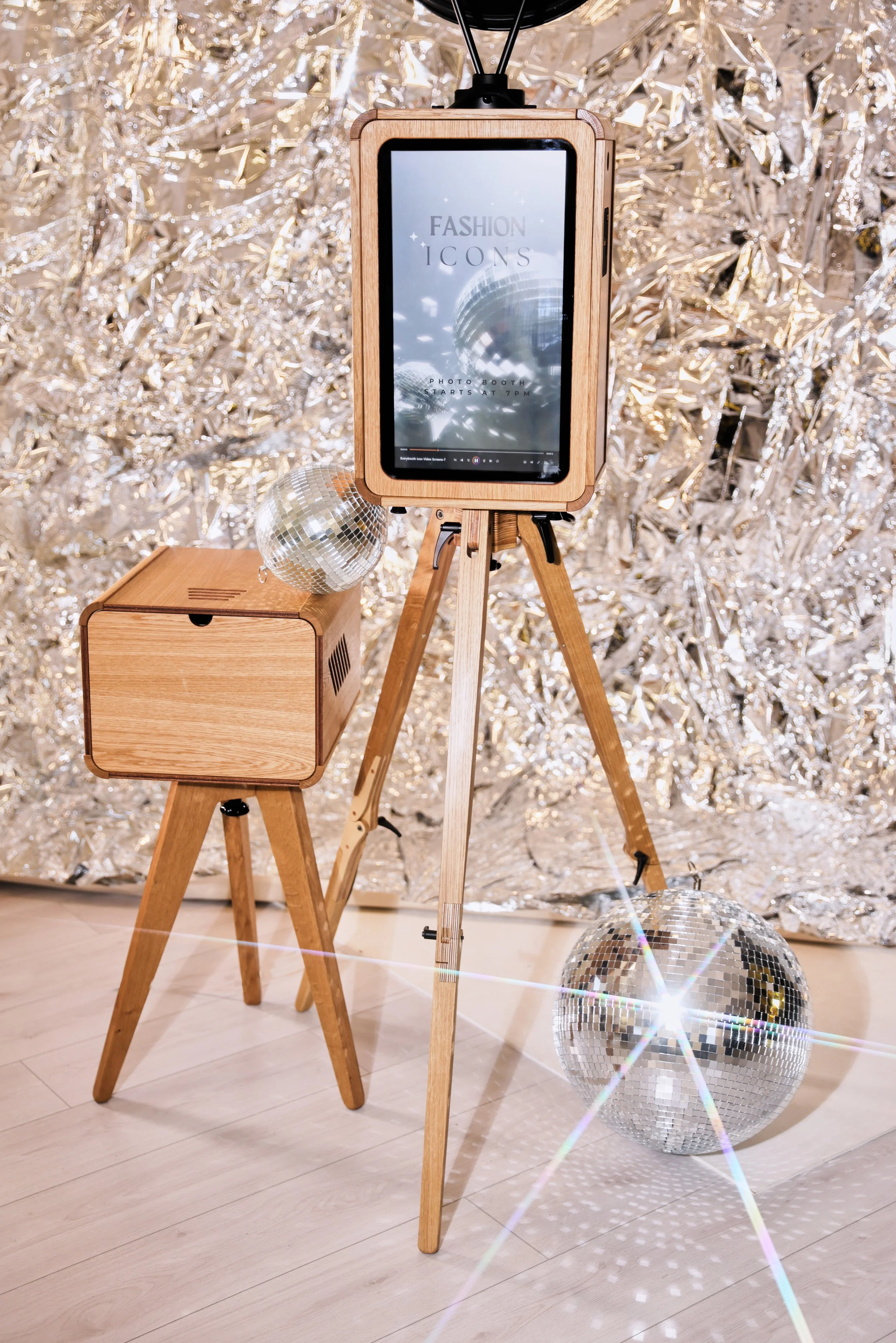 A wooden photo booth with a digital screen displaying the words 'Fashion Icons' and a description of the event, set up on a tripod. Two disco balls are on the floor, reflecting light, with silver foil decorations in the background.