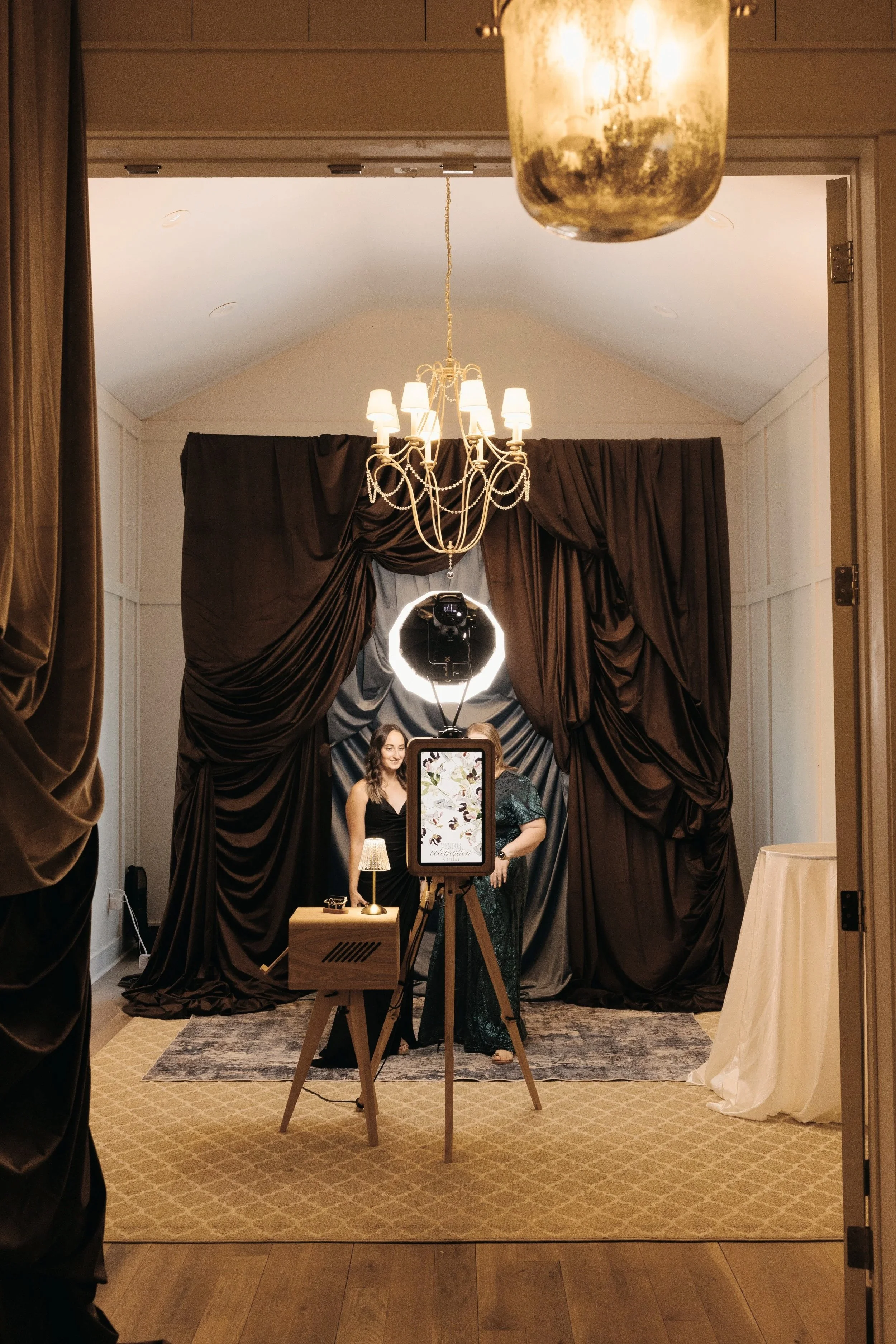 People taking photos in a photo booth with dark curtains, a chandelier, and a sign that reads 'Celebration' in a hotel conference room.