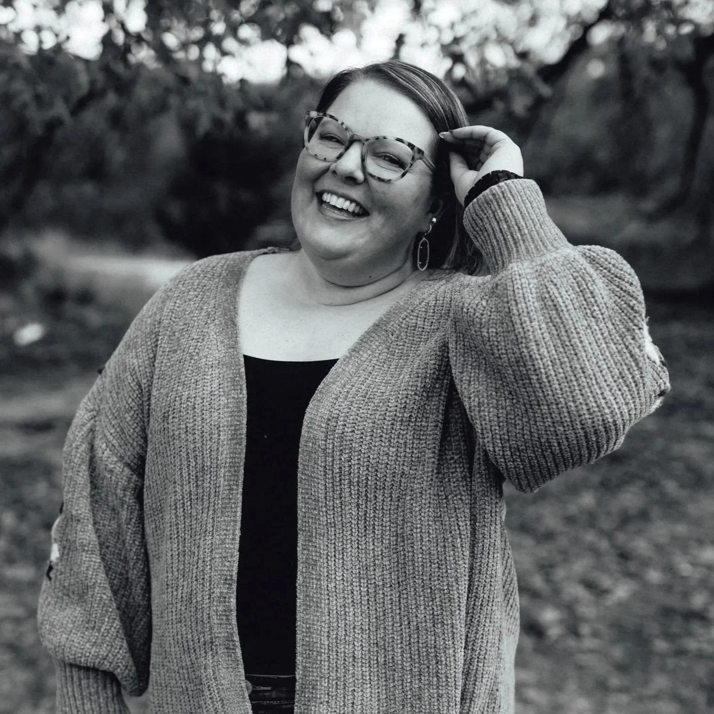 A woman smiling outdoors, wearing glasses, a black top, and a knitted cardigan. She has hoop earrings and a wristwatch, with her hand touching her hair. The background is blurred with trees.
