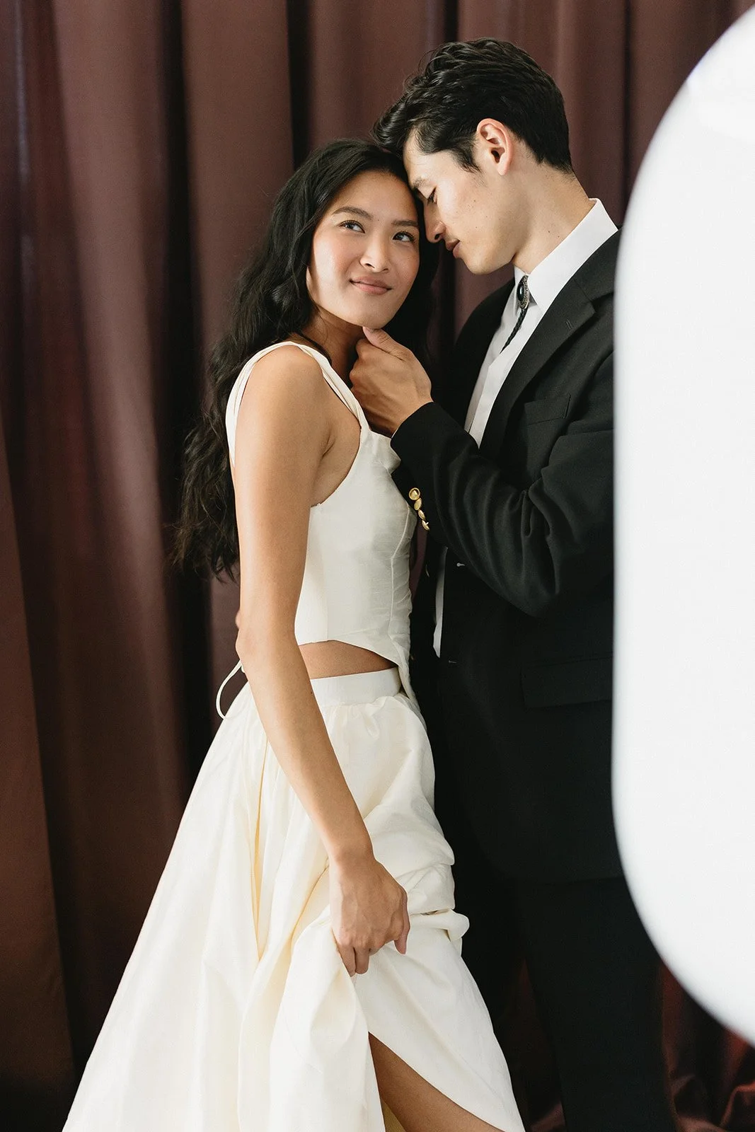 A couple standing close together, the man dressed in a black tuxedo and the woman in a white dress, with their foreheads touching in an intimate moment.