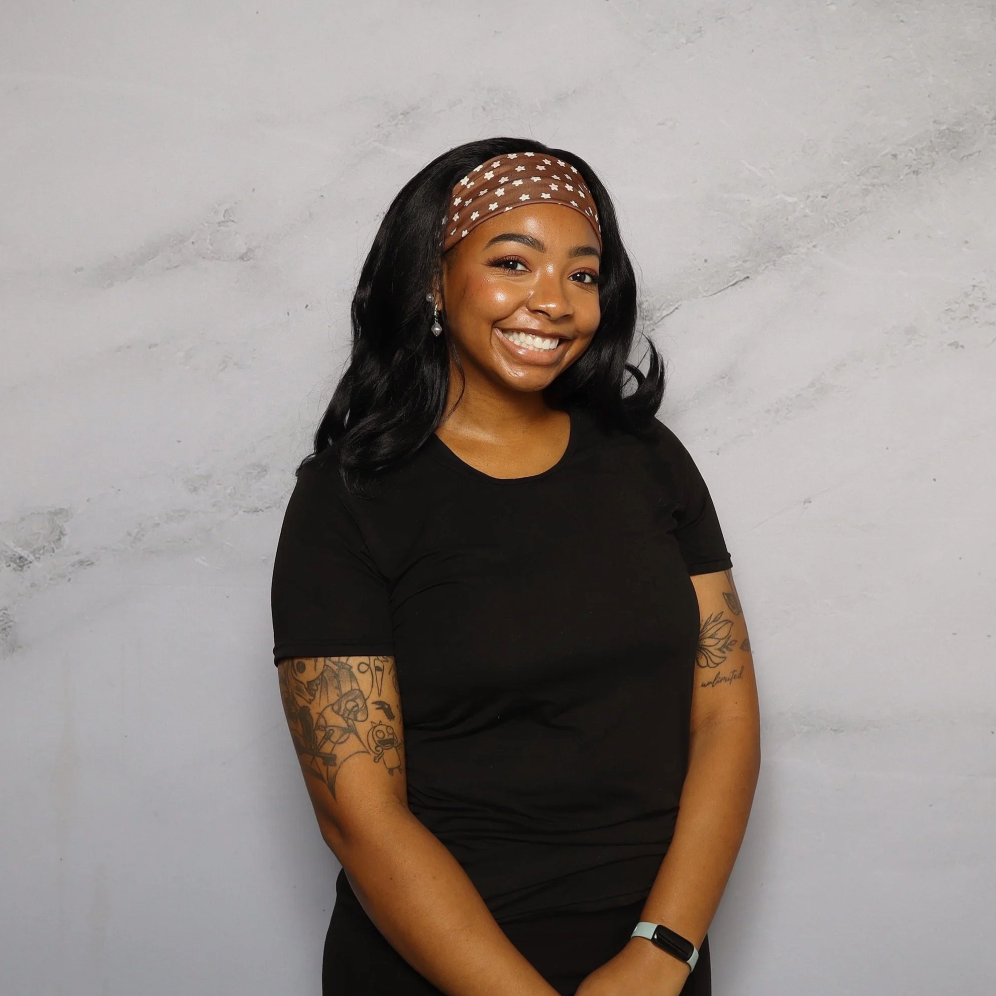 Smiling woman with long black hair wearing a brown headband with white floral pattern, black shirt, earrings, and a white wristband, standing against a light gray textured wall.