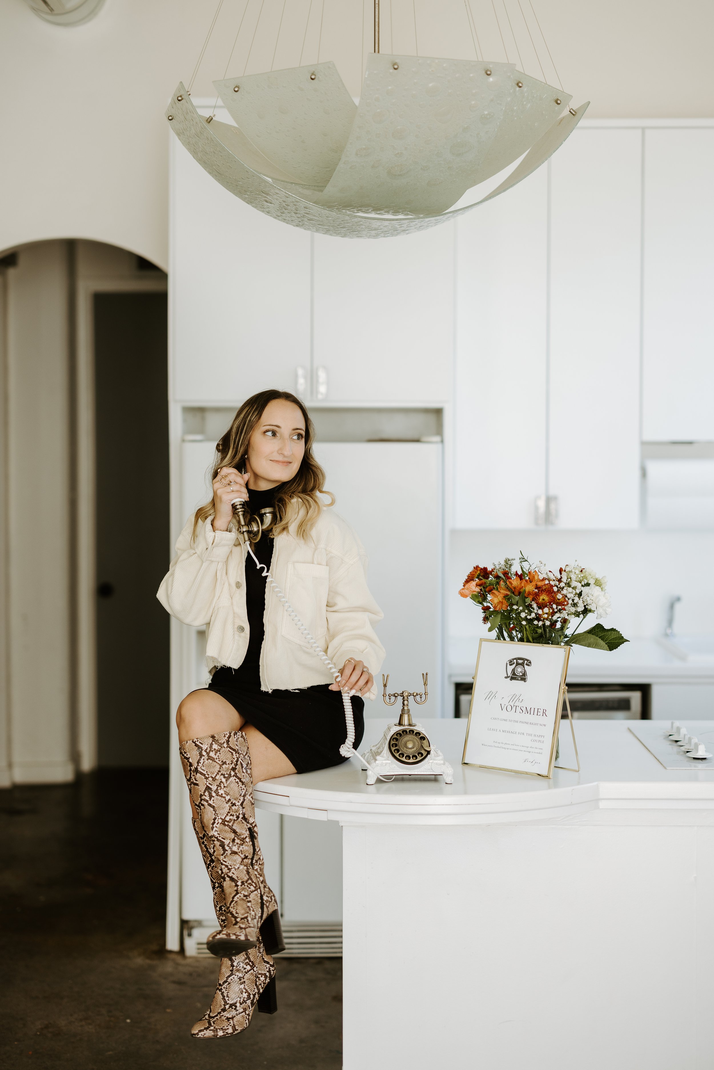 A woman sitting at a white table, talking on a vintage rotary telephone, with a bouquet of flowers, a framed sign, and decorative items on the table. She is wearing a black dress, a light-colored jacket, and snakeskin-patterned thigh-high boots, with her hair styled in loose waves.