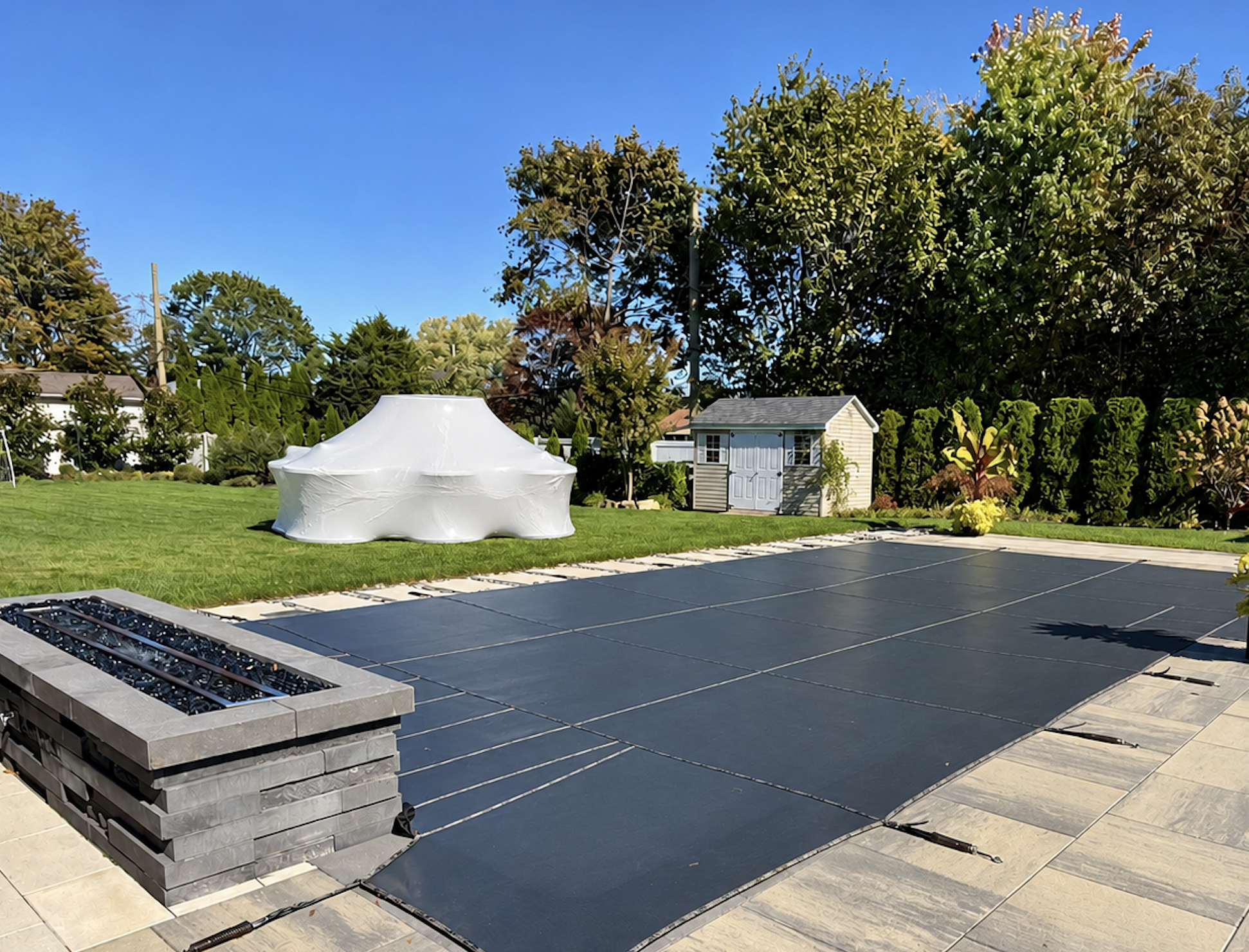 Sunlit backyard in Long Island with shrink wrapped furniture and a closed pool winterized on the ground, a small storage shed, lush trees, green grass, and a clear blue sky.