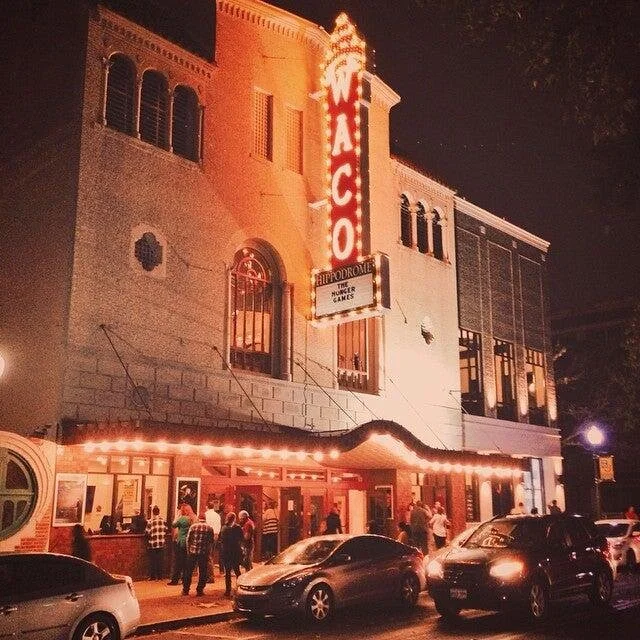 Nighttime view of a vintage theater with a large vertical neon sign that says 'WACO'. The building has an entrance with marquee lights and people standing outside.