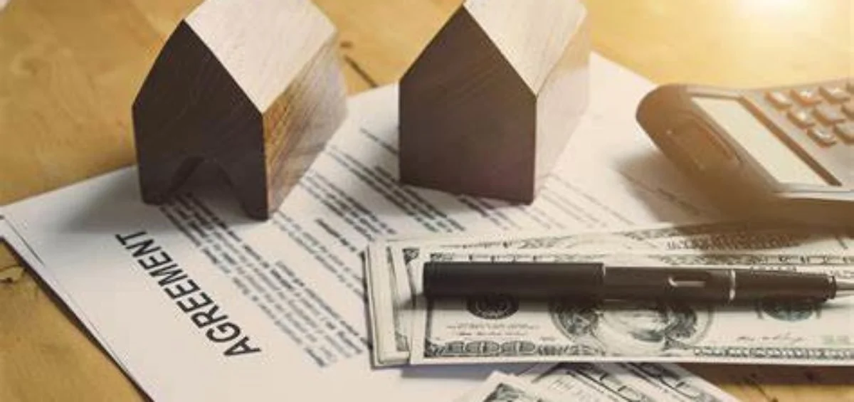 Two small wooden house models, a calculator, a pen, and U.S. dollar bills on real estate documents and an agreement sheet.