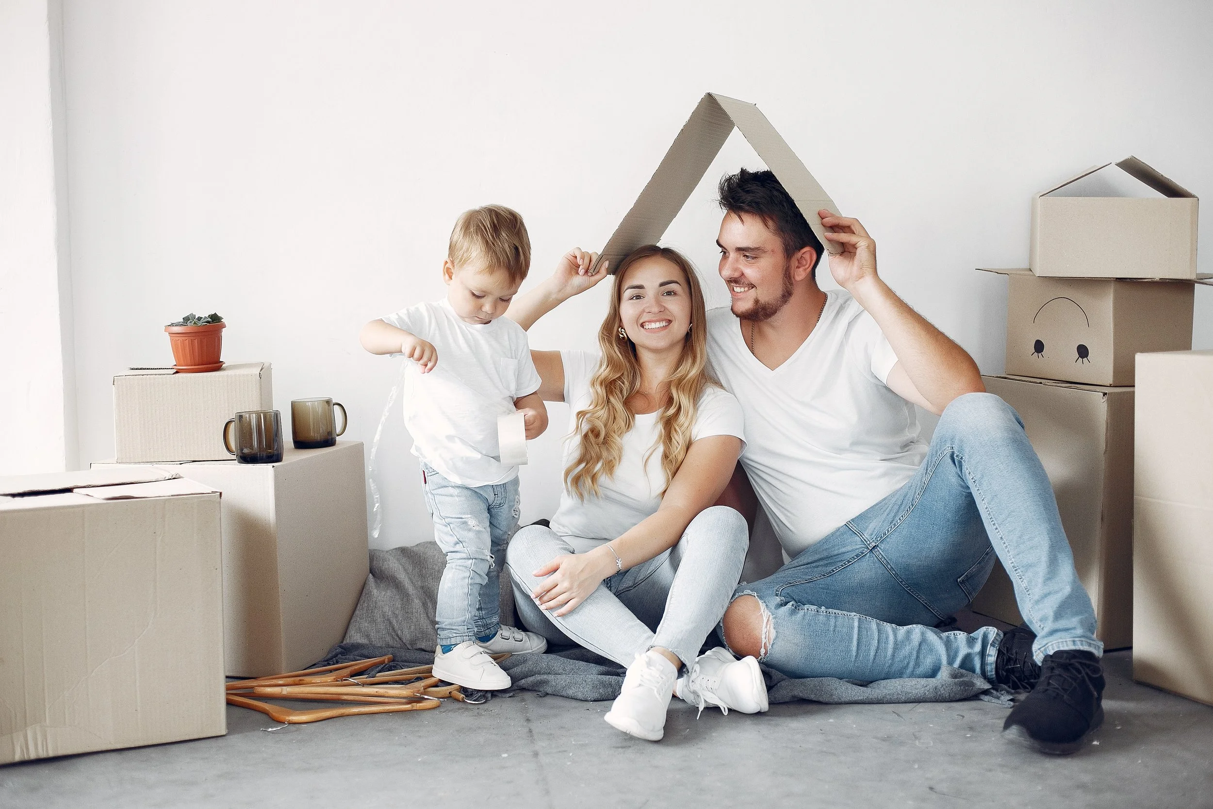 A family of three smiling and playing inside a room with moving boxes. The father and daughter are holding a cardboard piece above their heads, creating a house shape.