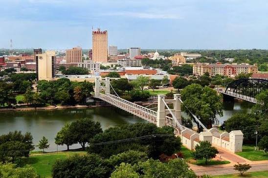 View of a city skyline with tall buildings, a river, and a suspension bridge connecting two parks.