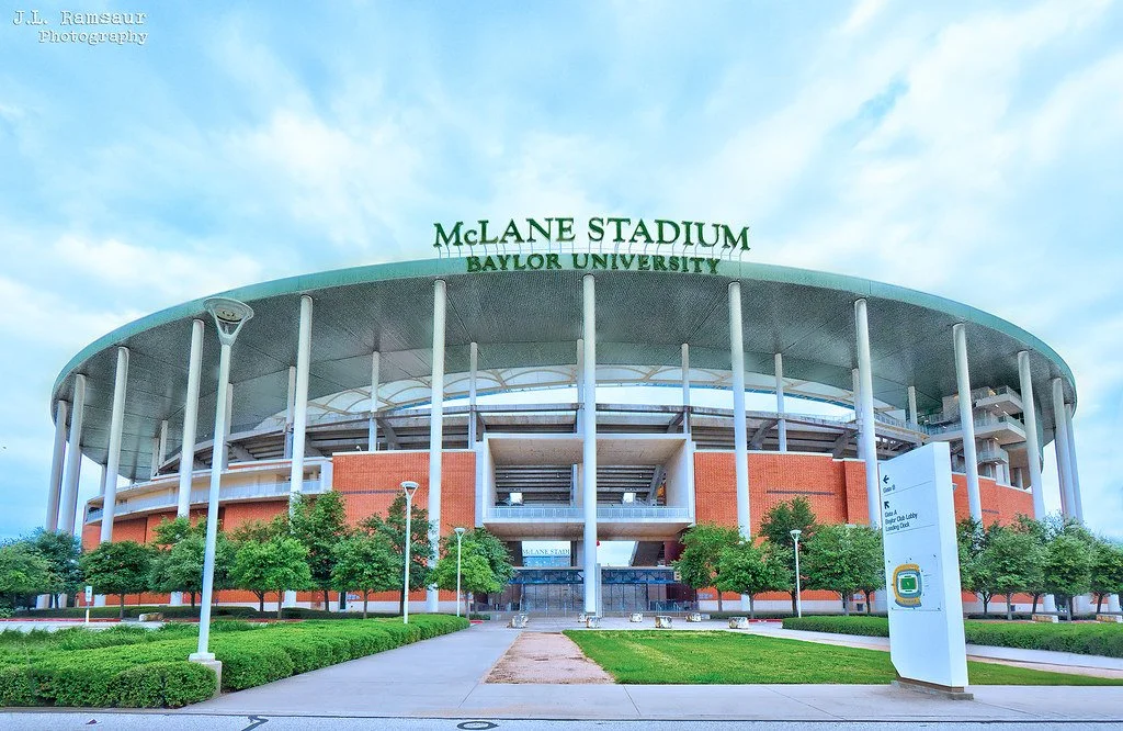 Front view of McLane Stadium at Baylor University, featuring a circular roof supported by tall white pillars, surrounded by green trees, with a pathway leading to the entrance and a sign in the foreground.