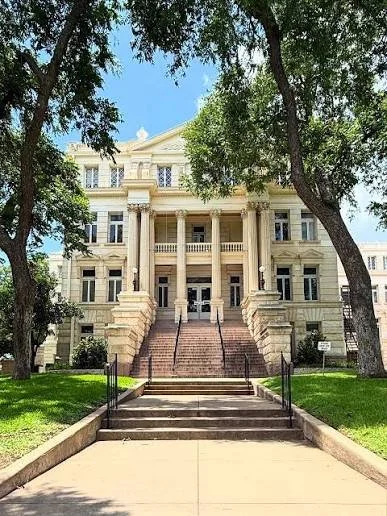 A grand neoclassical building with tall columns and a staircase leading to the entrance, surrounded by large trees and a well-maintained lawn.