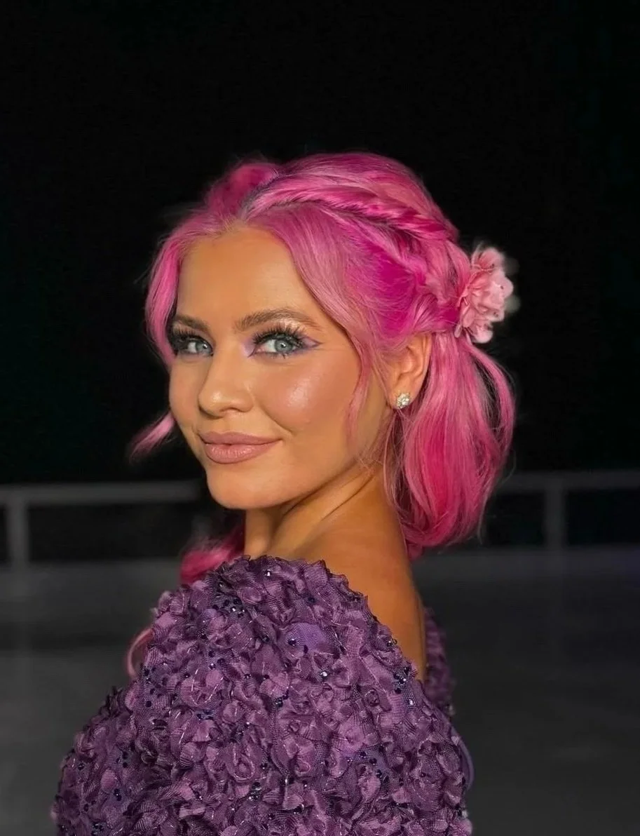 A woman with pink hair styled in loose waves and a braid, wearing a purple textured dress with embellishments, and small earrings, smiling against a dark background.