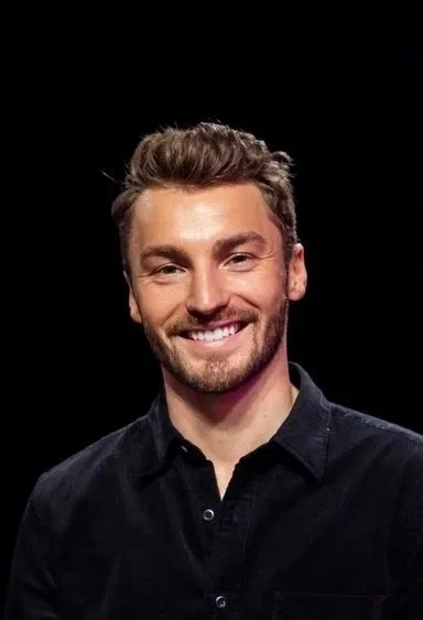 Portrait of a smiling young man with short, light brown hair and a beard, wearing a black button-up shirt against a black background.