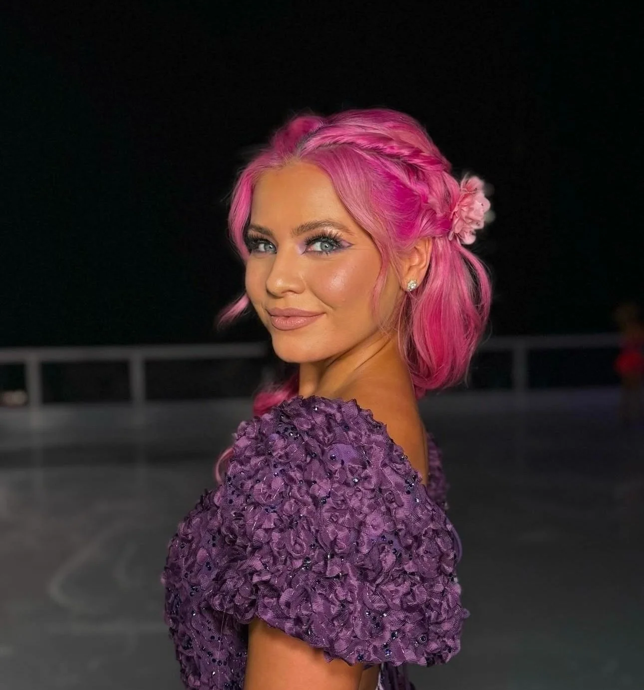 A woman with pink hair styled with braids and flowers, wearing a purple dress with textured fabric, smiling and looking at the camera at night.
