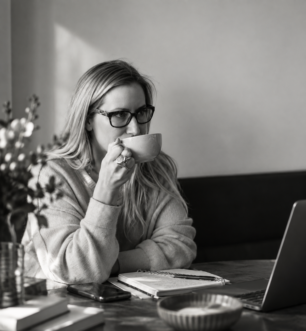 Black-and-white photo of a woman wearing glasses, holding a mug while working at her laptop.
