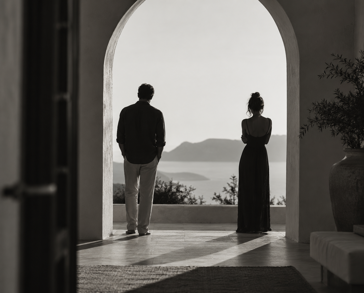 Black-and-white photo of two people standing apart on a balcony, looking toward a distant landscape of water and mountains.