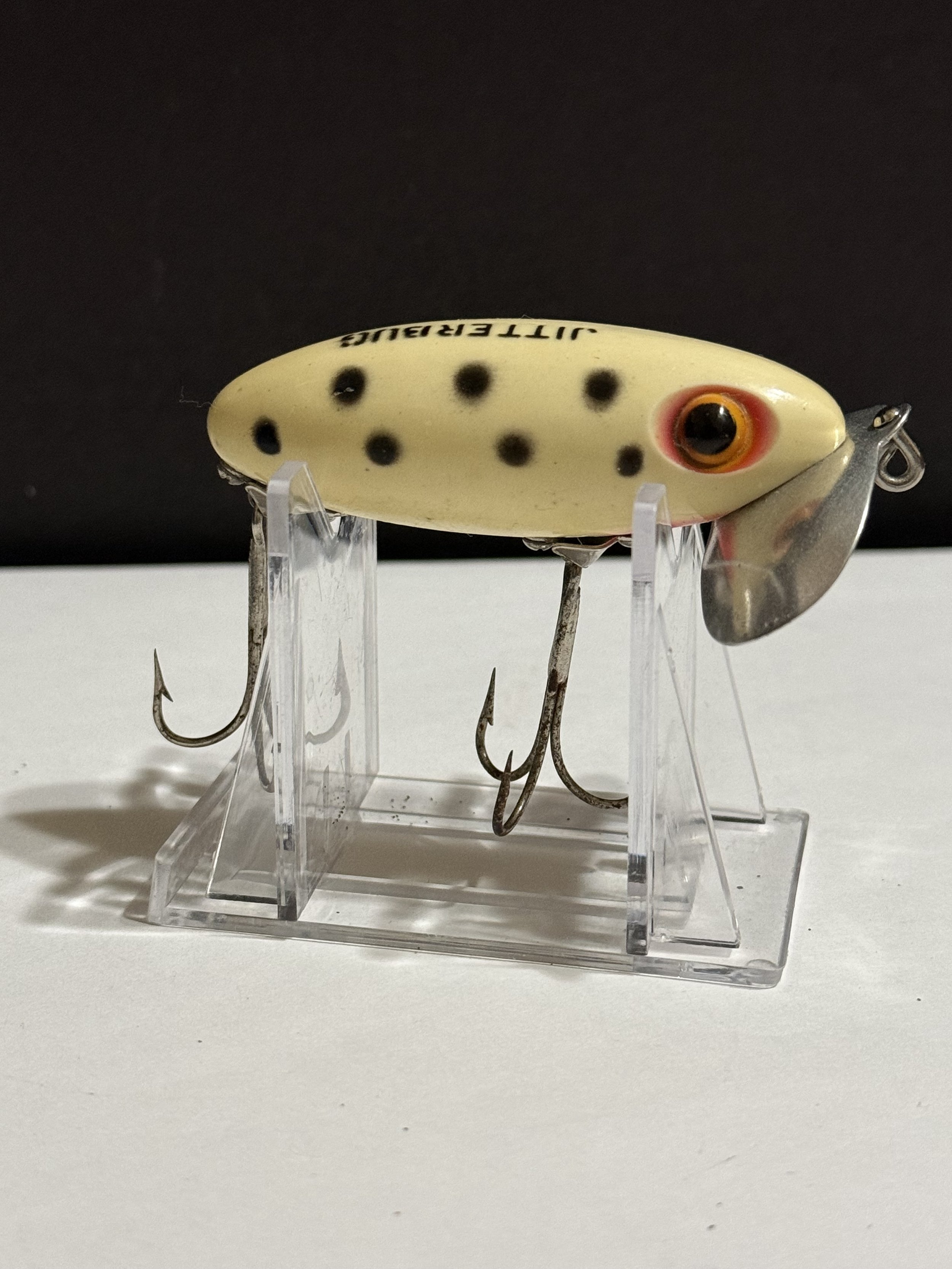 Close-up of a fishing lure with a yellow body, black spots, and a large orange and black eye, displayed on a clear plastic stand against a dark background.